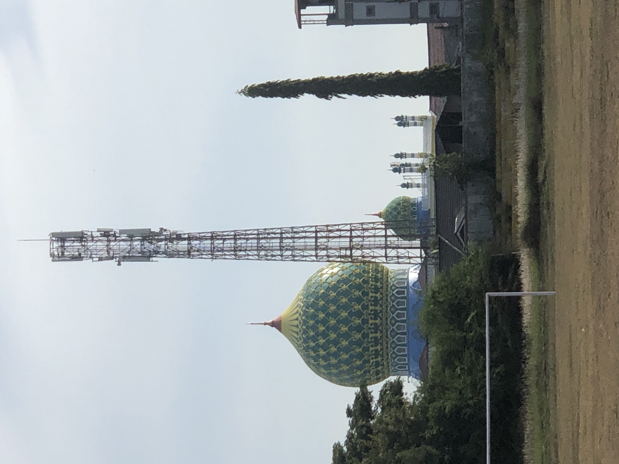 This image shows a wide shot of a mosque with a green and gold dome, a tall telecommunications tower, and a tall, narrow tree. In the foreground, a grassy field with a white soccer goal is visible. In the background, several smaller domes and minarets of the mosque can be seen, as well as some buildings and trees. The sky is clear and blue. The scene is likely located in Lhokseumawe, Indonesia, given the presence of Islamic architecture and the general environment.
