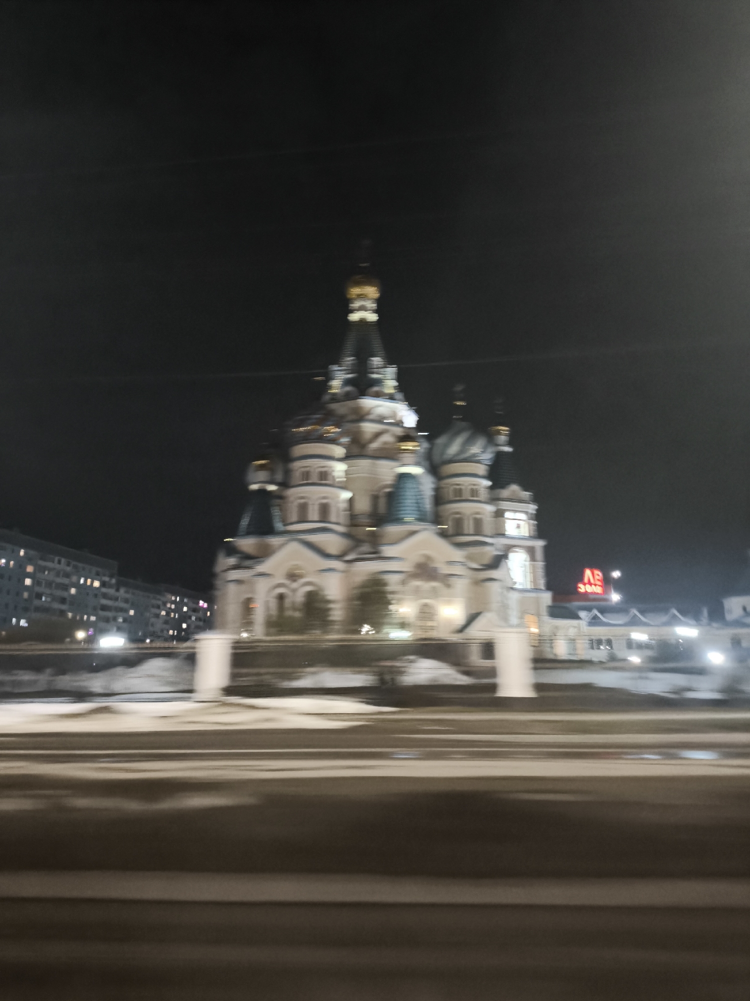 The image depicts a blurry night scene featuring the Alexander Nevsky Cathedral in Izhevsk, Russia. The cathedral, illuminated against the dark sky, is centrally positioned. In the background, other buildings are visible, including one with a red sign. The foreground shows blurred street details. No people are clearly discernible. The overall impression is of a nighttime view from a moving vehicle.