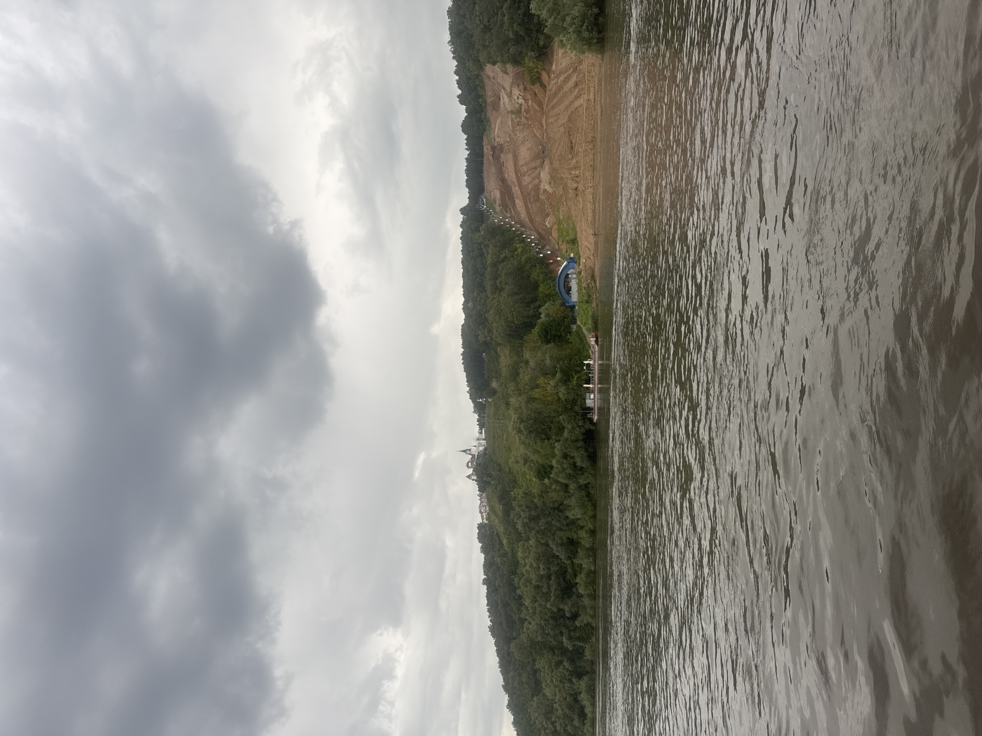 This image depicts a view from a body of water, likely a river, looking towards a hilly landscape under a cloudy sky. On the hillside, a forest covers much of the terrain. A section of the hill appears to have been cleared and reshaped, revealing bare earth with visible tire tracks, suggesting construction or landscaping work. A series of lights or poles are spaced out along a path leading up the cleared slope. At the base of the hill, near the water's edge, a small blue and white building with an arched entrance is visible, alongside a pontoon or small pier with a few people and what appears to be a small boat or barge. Further up the hill, nestled within the trees, is a cluster of white buildings with distinctive architectural features, possibly a church or monastery complex. The water in the foreground is choppy, reflecting the overcast sky. This scene is likely located in Ufa, Russia, given the presence of a large river and hilly terrain with architectural elements commonly found in the region.
