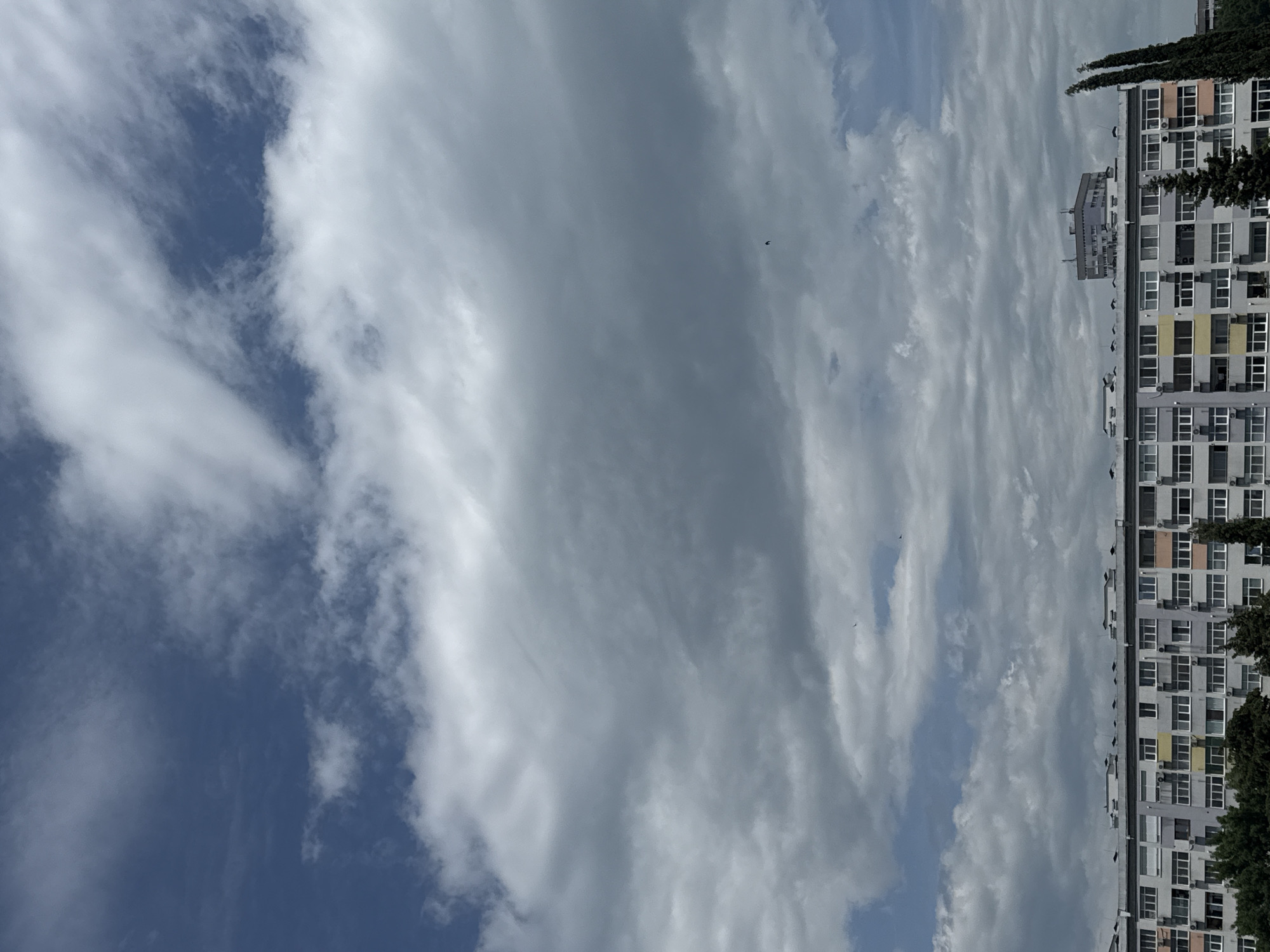 The image shows a high-angle view of a multi-story residential building in Burgas, Bulgaria. The building is light gray and features rows of windows, some with air conditioning units installed. Trees partially obscure the building. Above the building is a sky filled with layered clouds, ranging from white to gray. Two birds are visible in the sky.