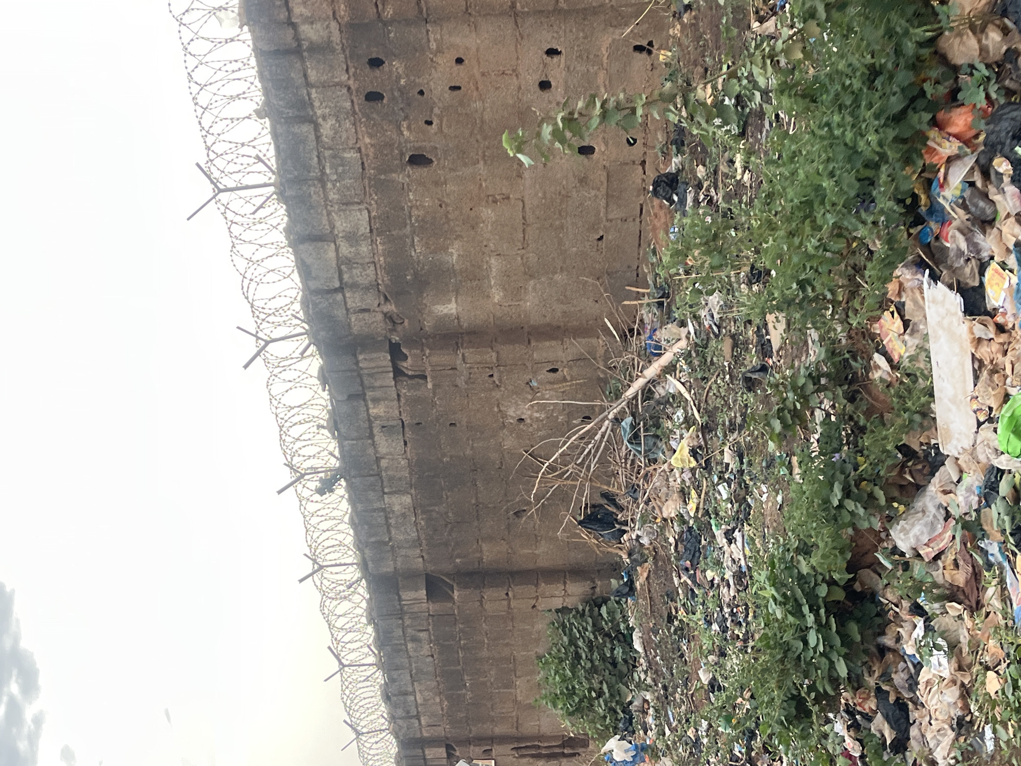 A high concrete wall topped with barbed wire separates a gray sky from a large accumulation of trash. The wall is pockmarked with holes. The garbage consists of plastic bags, bottles, and other discarded materials. Green plants sprout from within the trash pile. The location is likely Abuja, Nigeria, based on architectural and environmental cues.