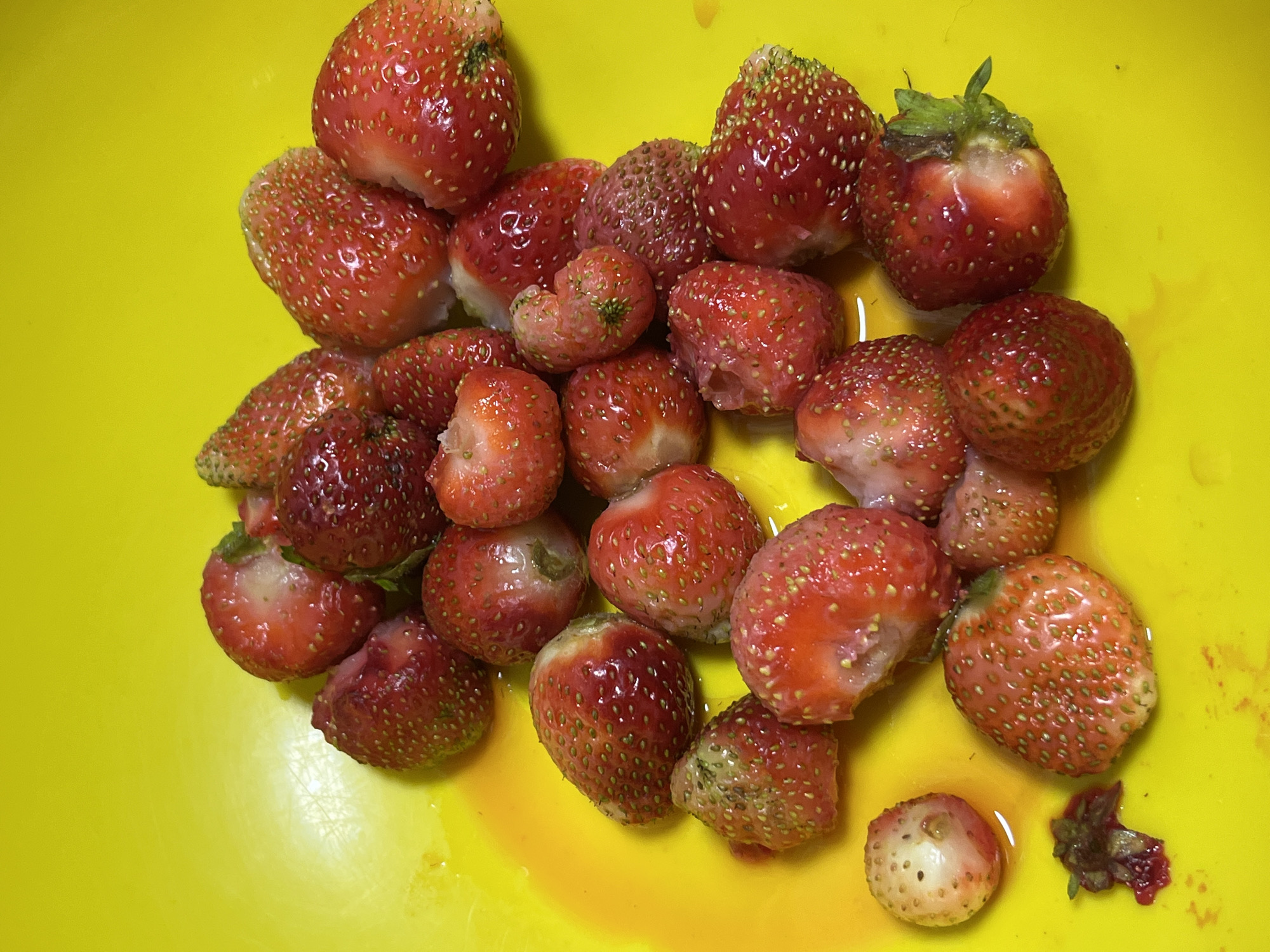 The image displays a pile of strawberries in a bright yellow bowl or plate. The strawberries are various shades of red, with some appearing juicier and more vibrant than others. Some strawberries are whole, while others appear to be cut or bitten into, revealing the white flesh inside. There are also some scattered seeds and what looks like a small piece of strawberry stem or leaf. The yellow surface is slightly wet, with some juice from the strawberries having seeped out. The lighting is somewhat uneven, casting shadows and highlighting the textures of the fruit. There are no people or other objects visible in the image, and there is no indication of the location in Nizhny Tagil, Russia.