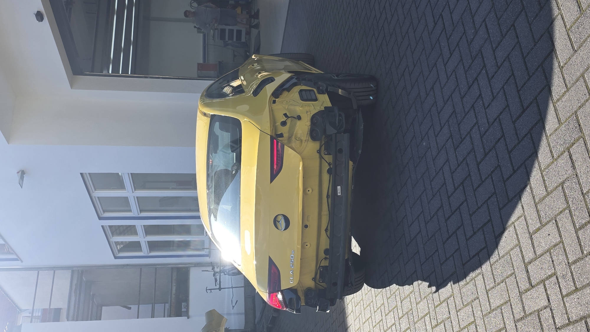 A bright yellow Mercedes-Benz CLA 250e is parked on a grey brick driveway in front of a white building with large windows. The rear bumper of the car is removed, and its taillights are visible. A person is standing inside the open garage door of the building, which is to the right of the car. The sunlight casts a shadow of the car onto the driveway.