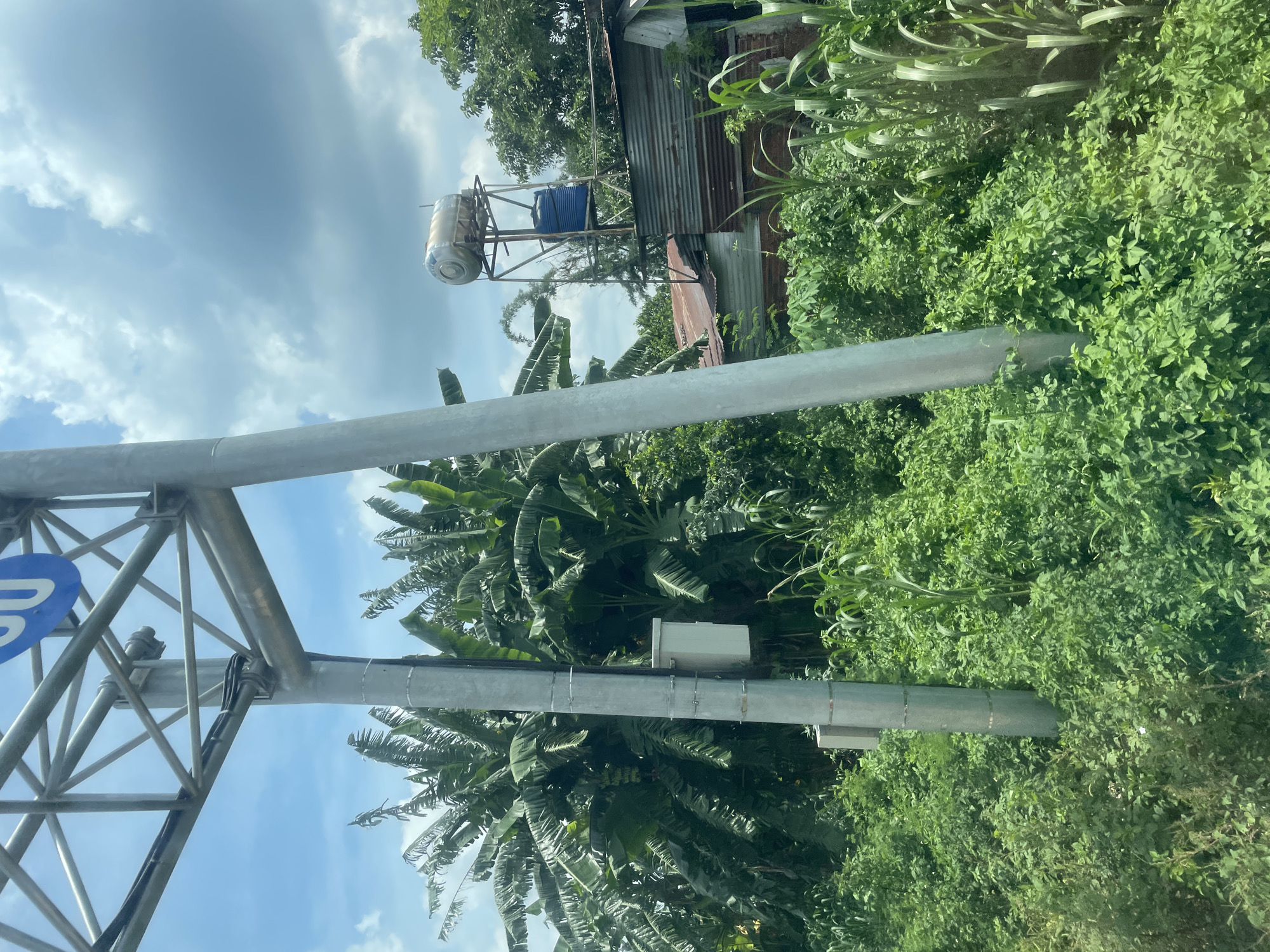 The image depicts a roadside scene with lush green vegetation, including banana trees and tall grass. A large metal support structure, likely for a sign, dominates the left side of the frame, with a blue circular sign with white lettering partially visible at the top. Several electrical conduits are attached to the support pole. To the right, a corrugated metal roof structure is visible, topped with a water heater and a blue plastic water tank on a metal frame. The sky is partly cloudy. There are no people visible in the image. The scene suggests a semi-rural or undeveloped area within Bình Xuân 2, Vietnam.