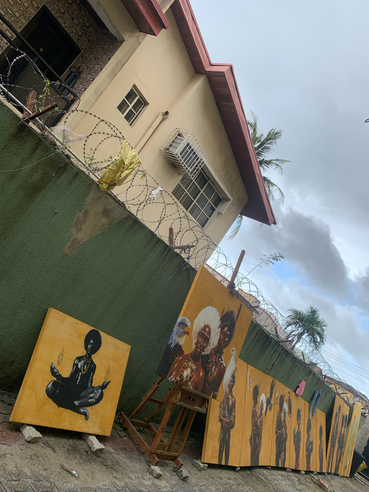 The image depicts a street scene in Ado, Nigeria, with a focus on artwork displayed outdoors. A cream-colored building with a reddish-brown roof is visible in the background, featuring barred windows and an air conditioning unit. A green wall, topped with barbed wire, runs along the foreground.
Leaning against the wall and on an easel are several large paintings on yellow canvases. The painting in the foreground shows a silhouetted figure in a meditative pose with flames emanating from their hands. To the right, another painting features portraits of people with elaborate hairstyles and an eagle and a dove. Further along the wall, a series of paintings depict groups of figures, some holding birds. Two pieces of clothing, one pink and one blue, are hanging from the barbed wire.
The ground appears to be paved with gray interlocking bricks, with scattered debris. The sky is overcast with scattered clouds and palm trees are visible in the background, suggesting a tropical environment. The overall impression is of an outdoor art display in a residential or commercial area of the city.