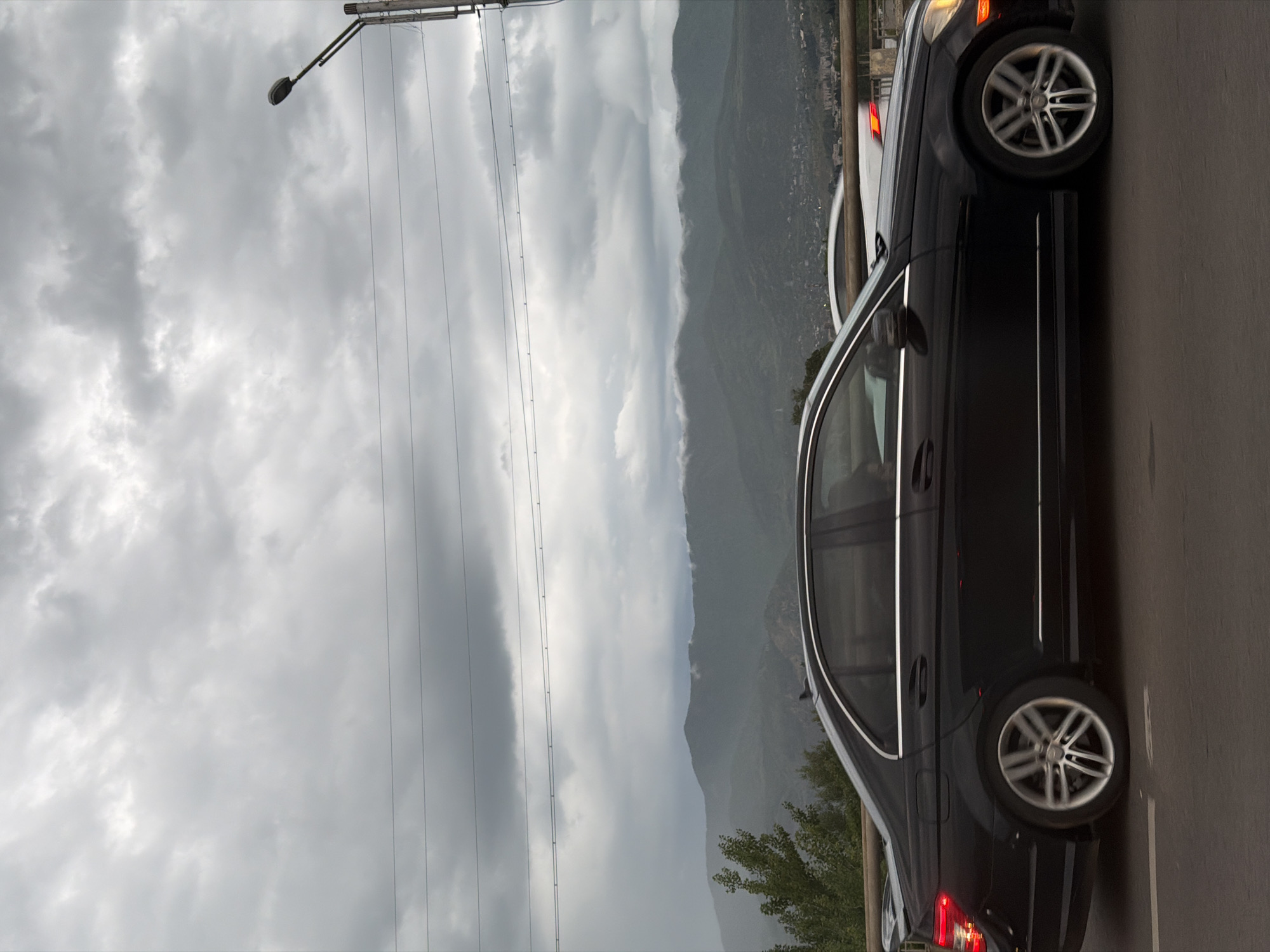 A dark-colored Mercedes-Benz sedan is on the right side of the road. The car's right tires and passenger side are visible. The background shows a cloudy sky, a utility pole, and mountains shrouded in mist. The city of Tbilisi, Georgia, is hinted at by the urban structures on the side of the mountain.