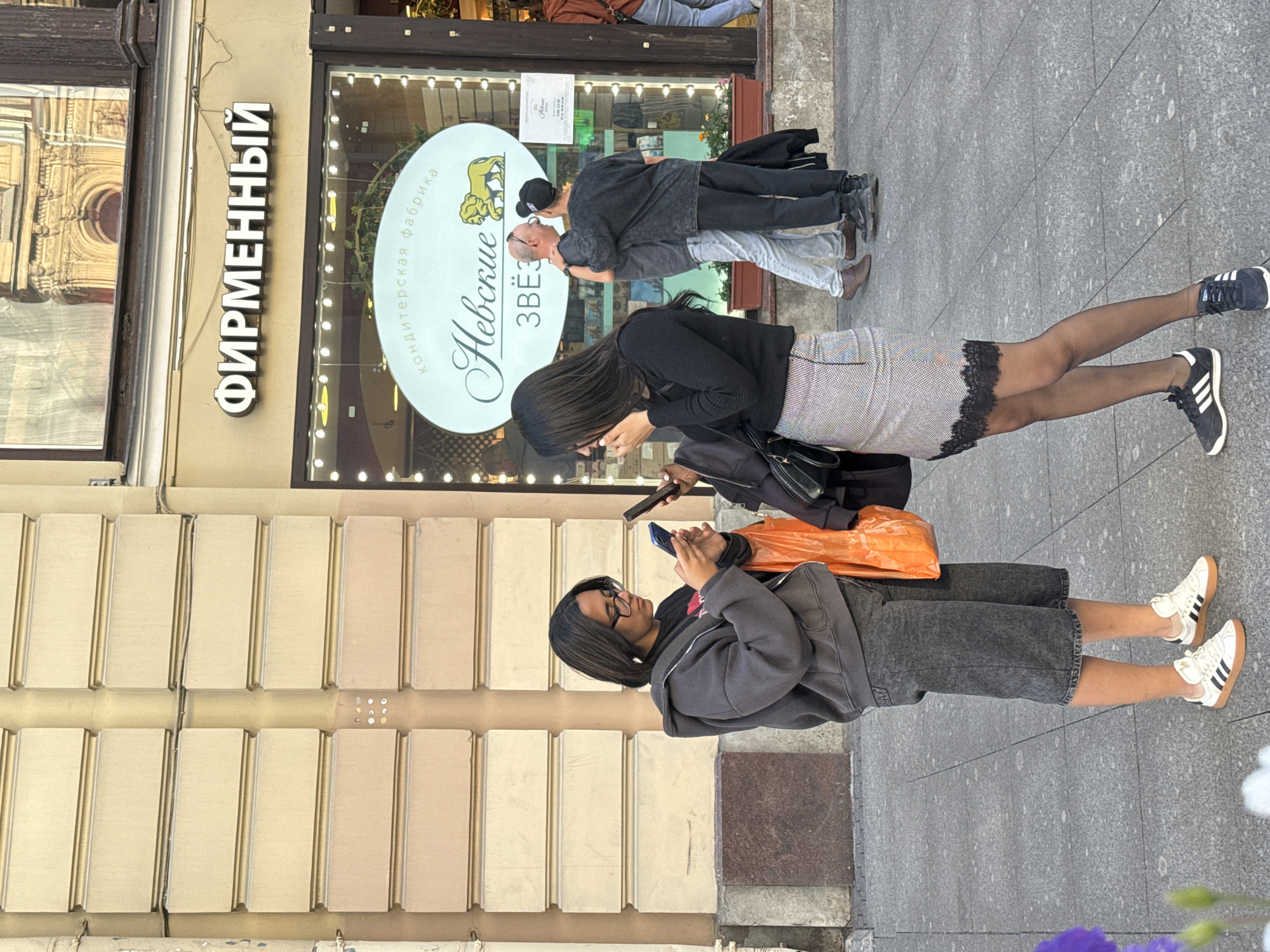 This media file shows a street scene in Saint Petersburg, Russia, with several people and a storefront. Two women are in the foreground, both looking at their phones. The woman closer to the camera is wearing glasses, a dark grey hoodie, and dark denim shorts. She has dark hair and is carrying an orange bag. The woman further away is wearing a black top, a textured skirt with black lace trim, and black tights with white-striped athletic shoes. Behind them, two men are standing close together, with one appearing to kiss the other on the cheek. They are both dressed in dark clothing.
The storefront behind the people features a prominent sign with white, illuminated letters that reads "ФИРМЕННЫЙ" (Firmenny), which translates to "Brand" or "Company." To the right of this sign is a circular logo with text in Russian, including "Невские Звёзды" (Nevskie Zvezdy), meaning "Nevsky Stars," and "кондитерская фабрика" (konditerskaya fabrika), indicating "confectionery factory." The facade of the building is made of light-colored rectangular panels. The ground is paved with grey tiles. The lighting suggests it is daytime, likely late afternoon or early evening, with soft, diffused sunlight. The overall atmosphere appears casual and urban.