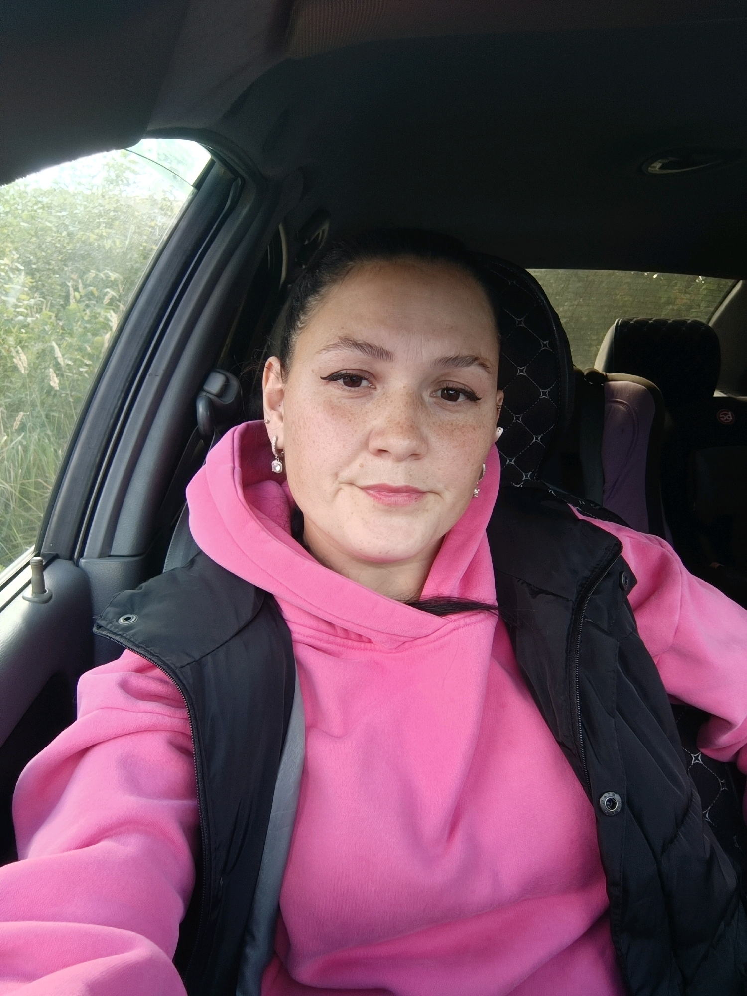 A woman with freckles and dark hair pulled back is seated in the driver's seat of a car. She is wearing a pink hoodie and a black vest. She is looking at the camera and smiling slightly. The car is parked outdoors, with greenery visible through the window. The image appears to have been taken during the daytime. The location is in Chaikovsky, Russia.
