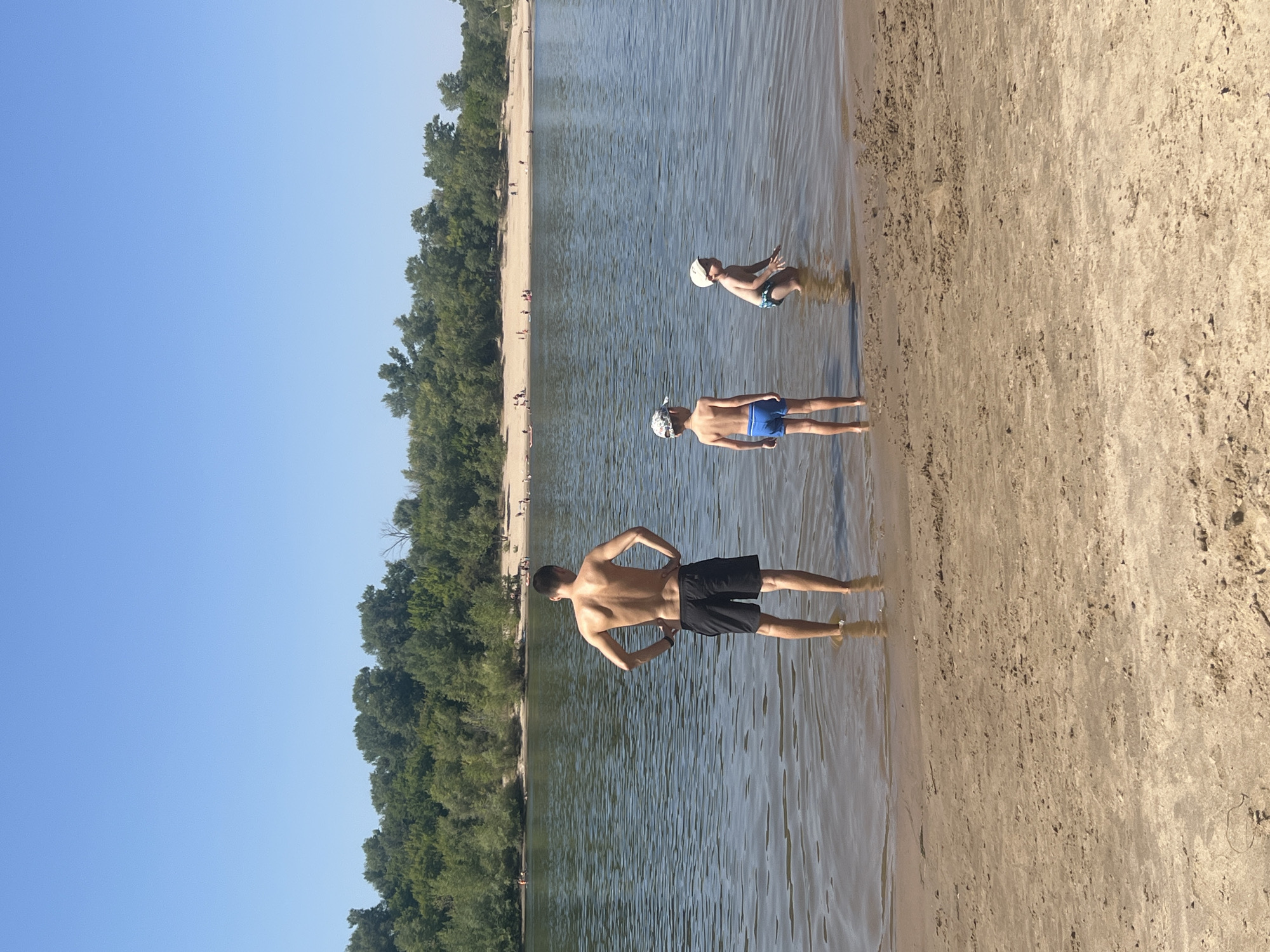 A man and two young boys are standing in shallow water on a sandy riverbank. The man, facing away from the camera, wears black shorts and has his hands on his hips. The two boys are also facing away from the camera. The boy on the left wears blue swim trunks and a patterned cap. The boy on the right wears patterned swim trunks and a white cap. They are on the bank of a wide, calm river with a tree-lined shore in the background. The sky is clear and blue. There are other people visible in the distance on the opposite bank, which appears to be a sandy beach with trees. The scene is set in the city of Kilyakovka, Russia, during the daytime.