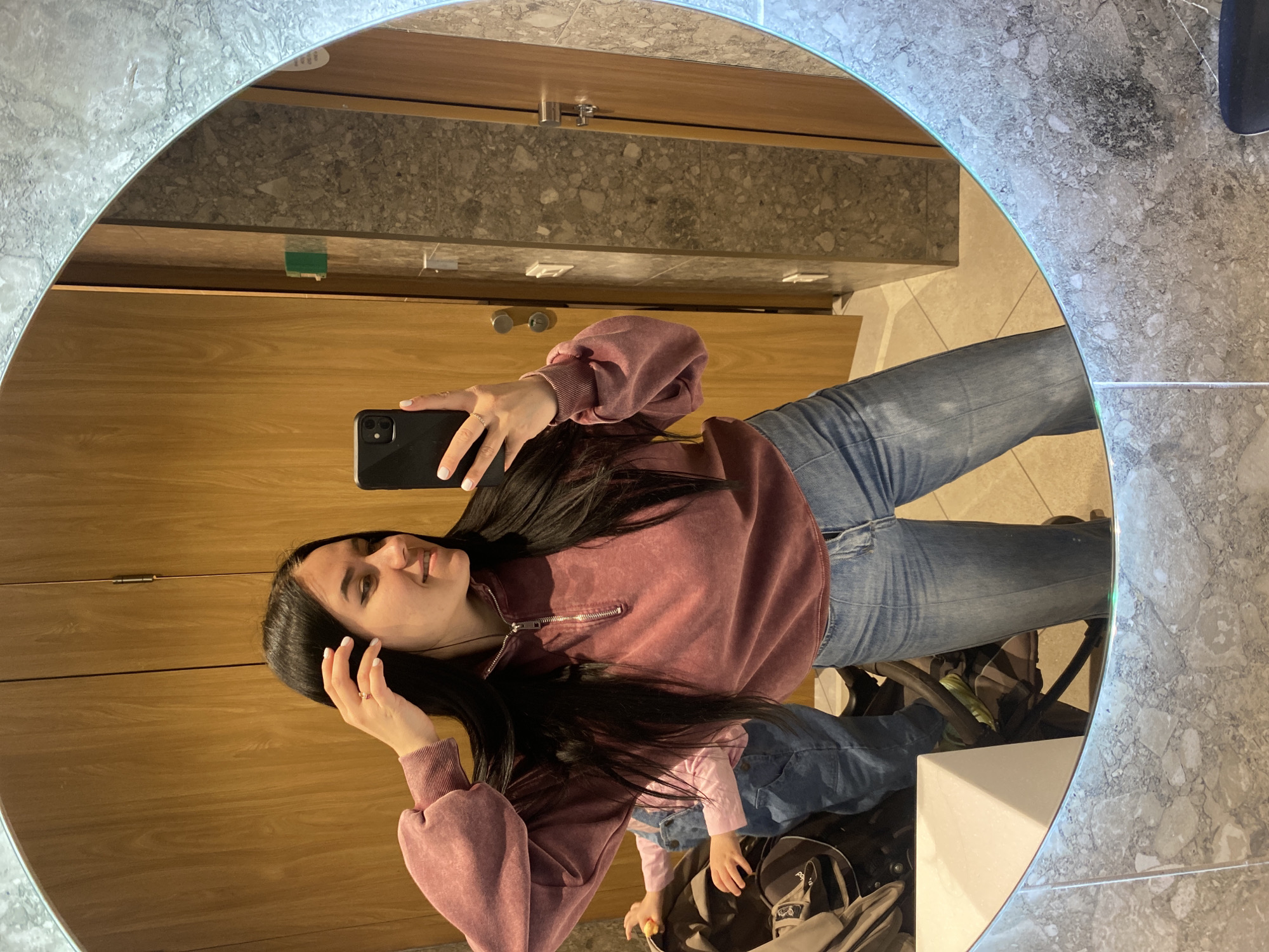 A woman is taking a selfie in a public restroom mirror. The mirror is round and has an illuminated edge. The restroom has light wood paneling and grey countertops and walls. The woman is wearing a mauve sweater and jeans. A young child in denim overalls stands near her legs. The location is potentially in the city of Perm, Russia.