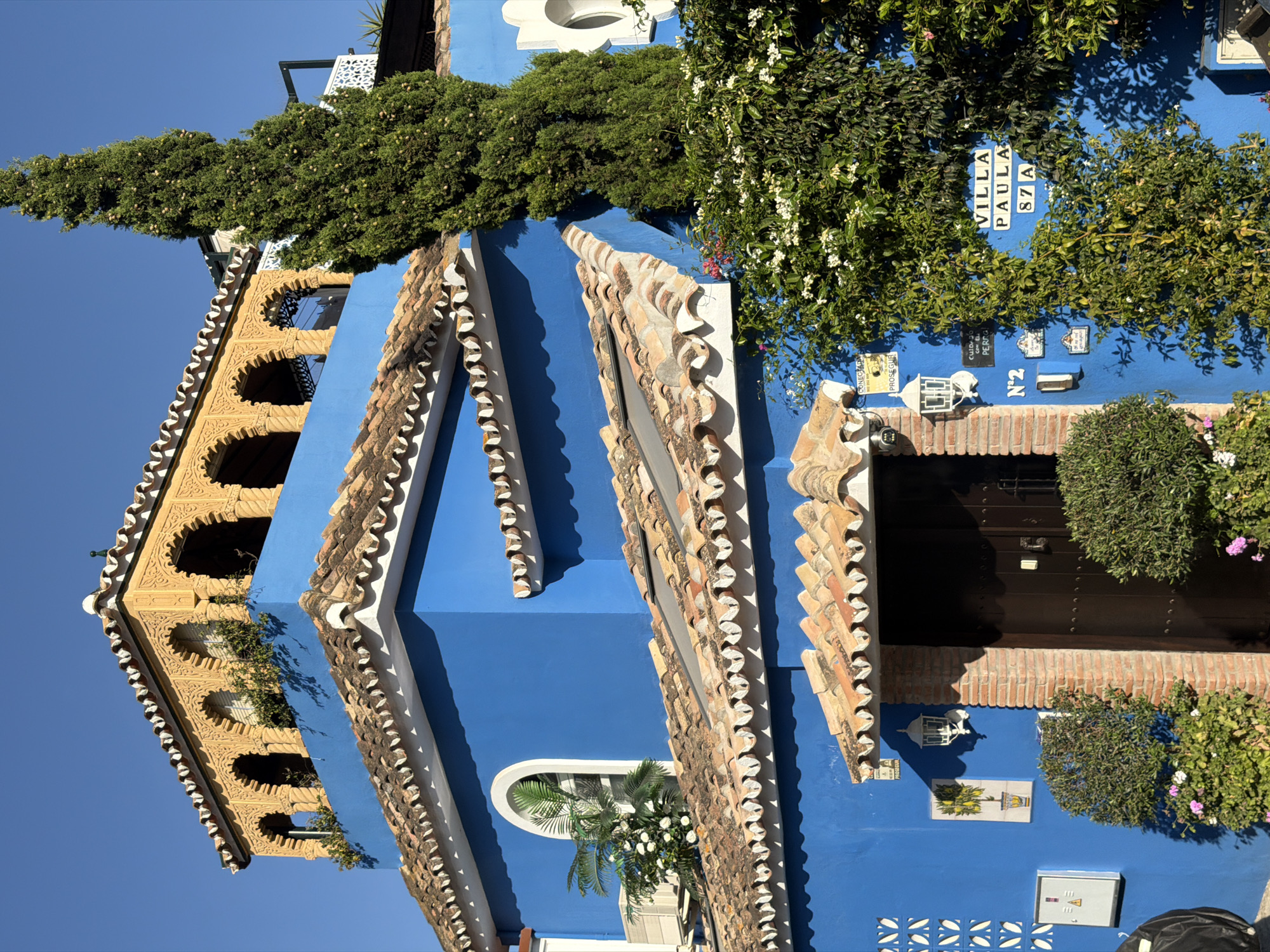 The image depicts the exterior of a vibrant blue villa, identified by signage as "VILLA PAULA 87A". The villa has a traditional Spanish architectural style, characterized by a terracotta tile roof with decorative white scalloped edging. The building features ornate, arched windows with yellow detailing on the uppermost level, resembling a Moroccan or Moorish influence. A tall, slender evergreen tree stands prominently to the right of the villa. Lush green foliage, including flowering vines with white blossoms, cascades around the entrance and parts of the villa's facade. The entrance itself is framed by exposed brickwork and a dark wooden door. Several small signs and a light fixture are visible near the entrance. The environment suggests a sunny day with a clear blue sky. The location is specified as Saladillo-Benamara, Spain. There are no people visible in the image.