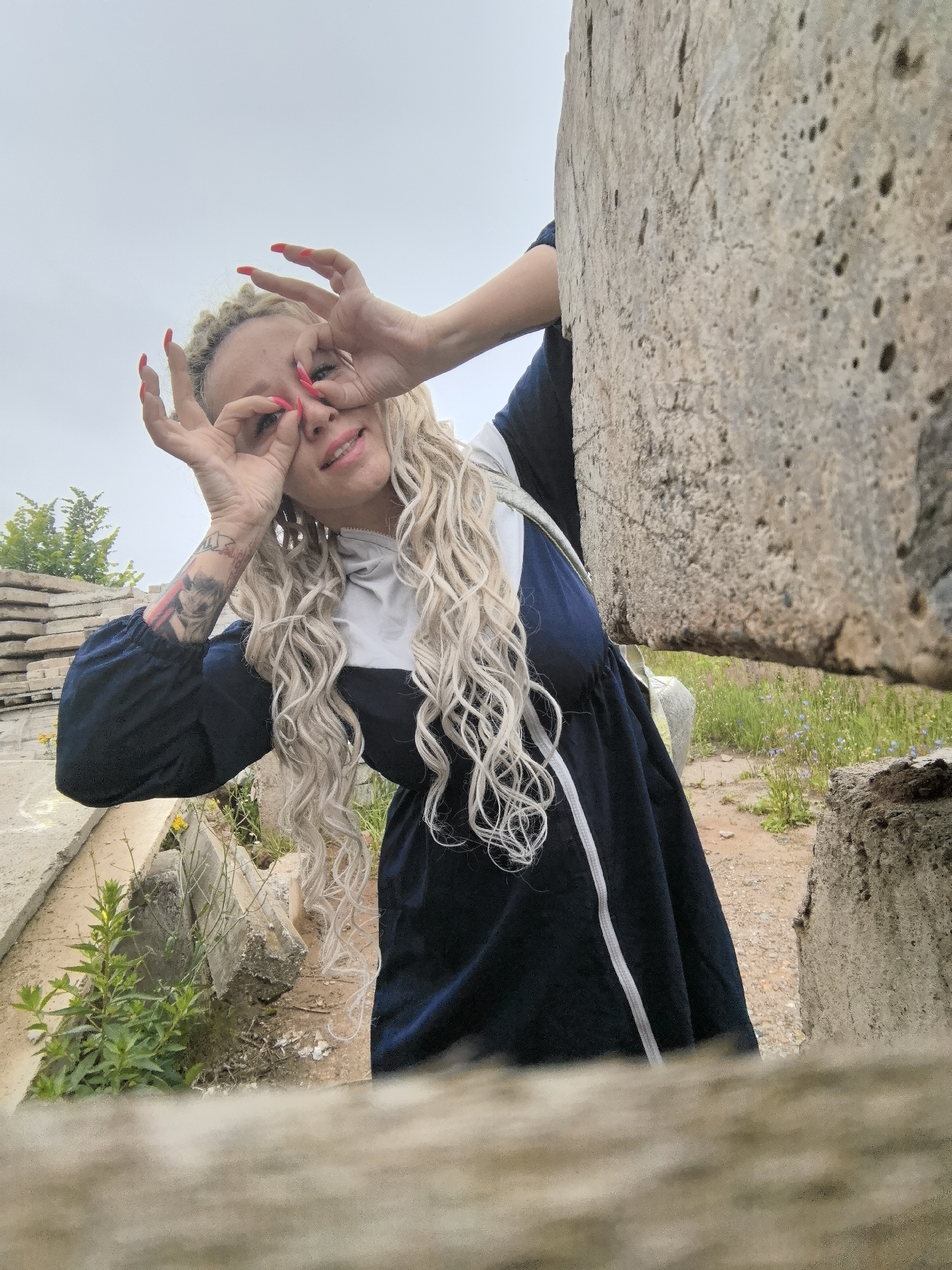 A woman with blonde, curly hair and bright red fingernails poses with her hands over her eyes, making a "binoculars" gesture. She wears a dark blue dress with white trim. The background suggests an outdoor, somewhat neglected area with concrete structures and vegetation. The sky is overcast. The scene is likely in Chaikovsky, Russia, given the context of the prompt.