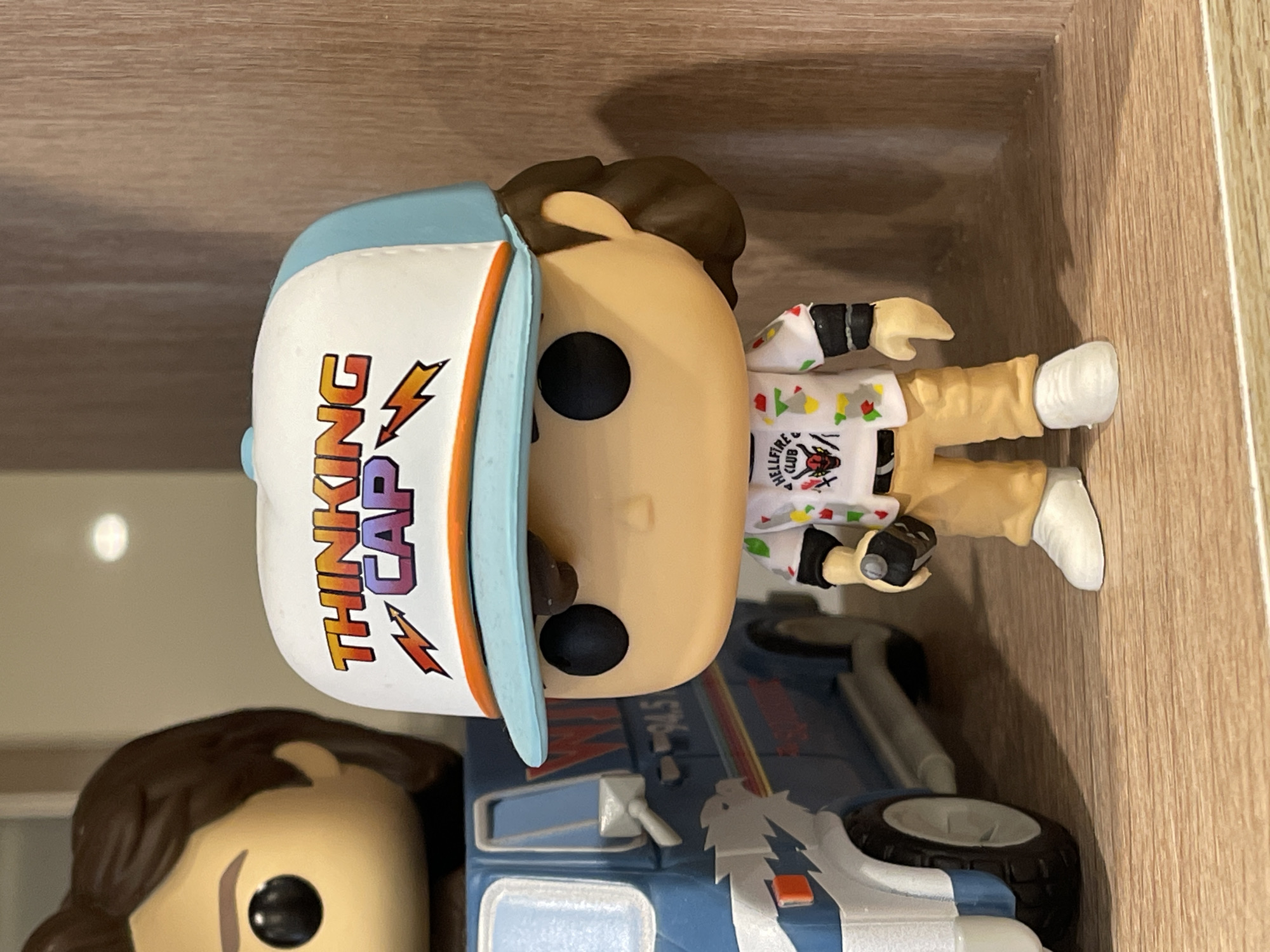 A close-up, eye-level shot features two Funko Pop figures and a toy van on a wooden shelf. The figure in the foreground is a detailed representation of Dustin Henderson from "Stranger Things," wearing a white cap with "THINKING CAP" in colorful letters, a short-sleeved shirt with a "HELLFIRE CLUB" logo and colorful graphics, tan pants, and white sneakers. He holds a black object in his right hand. To his left, partially visible, is another Funko Pop figure with brown hair and dark eyes. Behind Dustin is a blue toy van with the number "94.5" and graphics of a bird with lightning bolts. The background consists of a wooden surface. The image was taken in Khimki, Russia.