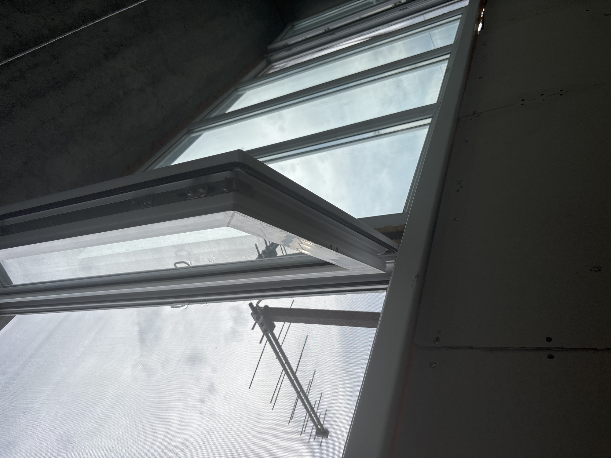 The image shows a low-angle view of an open window with a view of the sky and an antenna. The window appears to be a casement window, opening outwards from the bottom. The frame of the window is white, and there is a screen visible through the opening. The sky is overcast, with diffused light. An outdoor television antenna, a directional Yagi-Uda type, is visible outside the window, its elements extending horizontally. The antenna is mounted on a wooden crossbar which is attached to the building.
The interior framing of the window and adjacent wall are also visible. The wall appears to be drywall with visible seams and screw holes. Above the window, a concrete ceiling or overhang is visible, with a rough texture. The scene suggests an indoor setting looking out at the sky and external structures. The lighting indicates it is daytime, likely overcast due to the diffused light and cloud cover. There are no people visible in the image. The overall mood is neutral, focusing on the architectural and environmental elements. No text is visible in the image.