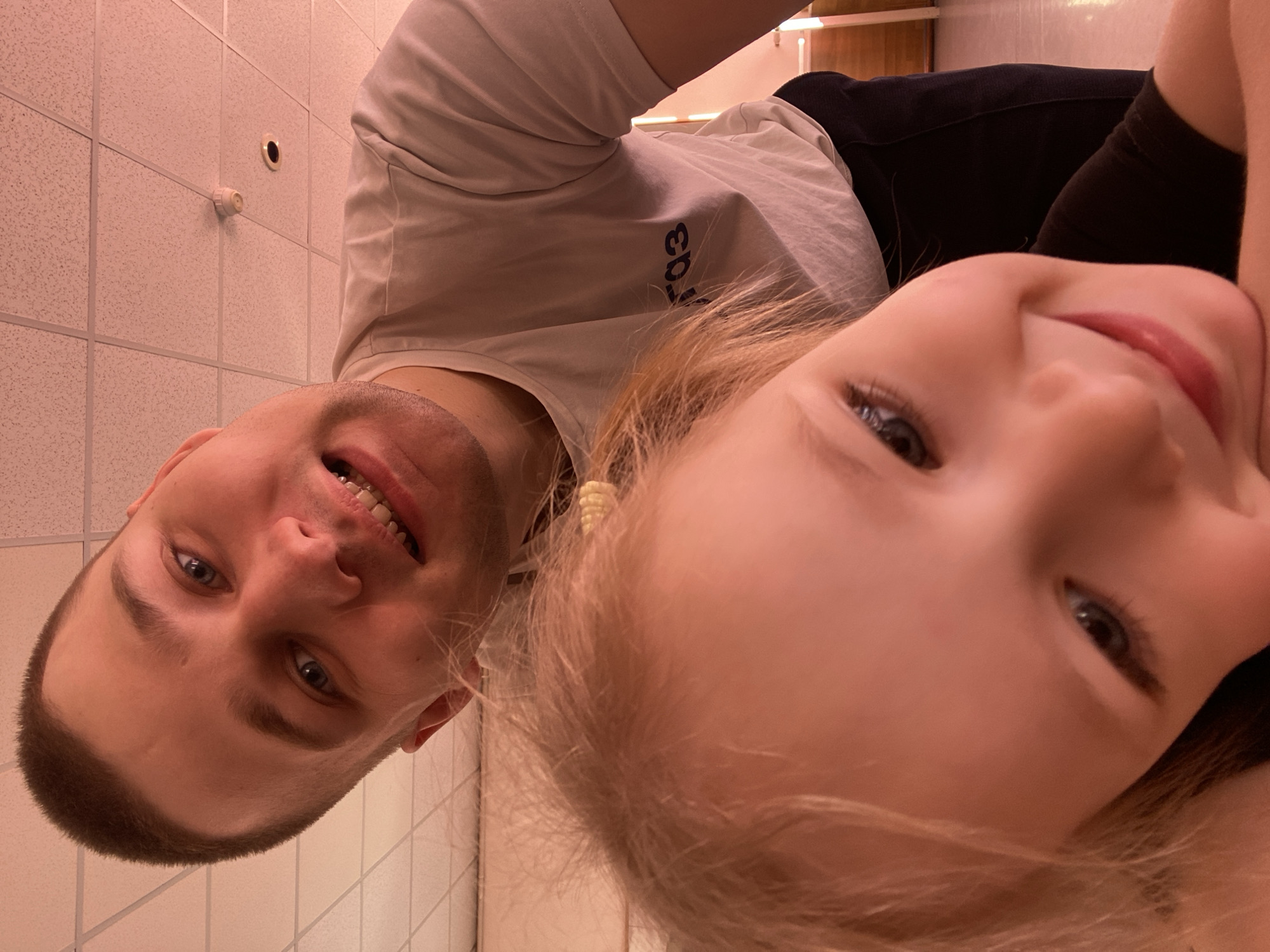 A selfie-style, close-up image depicts a man and a young girl. The man, on the left, has short brown hair, blue eyes, and is wearing a light-colored t-shirt. The girl, positioned closer to the camera on the right, has blonde hair and light skin. The background shows a tiled wall and ceiling, possibly indicating a bathroom or similar institutional setting.
The image was taken in the city of Chaikovsky, Russia.
