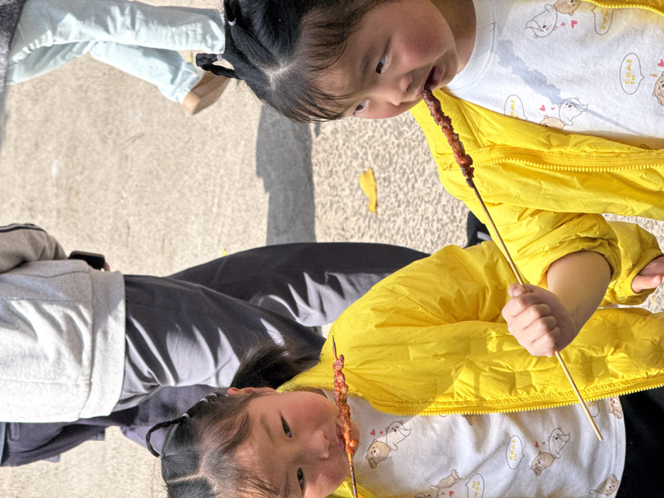 Two young East Asian children, dressed in matching bright yellow puffer jackets over white t-shirts patterned with cartoon dogs, are depicted outdoors in Jiulongpo District, China. The child on the left, with dark hair styled in two pigtails, actively eats a reddish-brown food item from a wooden skewer, with residue visible around her mouth, while looking towards the viewer. The child on the right, also with dark hair in pigtails, holds a similar wooden skewer in her right hand and bites into the food, looking towards the right side of the frame. In the blurred background, partial views of several adults are visible, including one wearing grey pants and a light grey sweatshirt, carrying a dark object, and another in light blue pants with brown shoes. The subjects are positioned on a light-colored paved surface, likely concrete or asphalt, which displays strong shadows indicating direct sunlight. A few scattered yellow leaves are present on the ground.