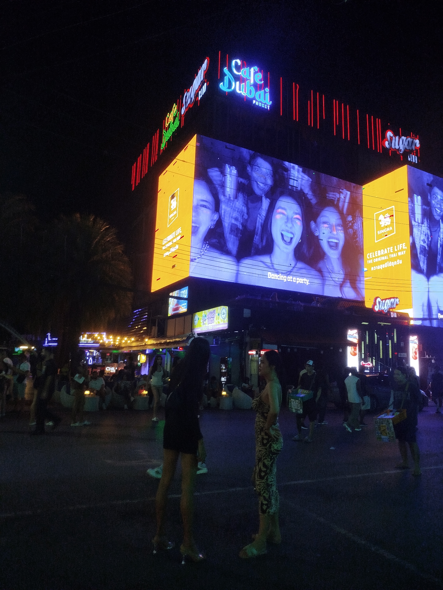 This is a nighttime outdoor shot of a street scene with prominent neon signage. A large, brightly lit billboard displays images of people's faces with glowing eyes and the text "CELEBRATE LIFE, THE ORIGINAL THAI WAY" in English and Thai. Above this billboard, neon signs read "Cafe Dubai Phuket" and "Sugar CLUB". The street is populated with people, some of whom are walking or standing. In the foreground, two women are visible. One woman on the left is wearing a patterned dress and standing to the side. The other woman on the right is wearing a black dress and appears to be walking away from the camera. The street is dark, with artificial lighting from the signs and some scattered lights from nearby establishments creating a vibrant, nocturnal atmosphere.