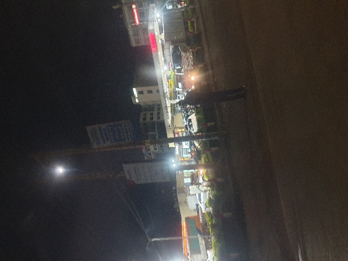 This media file captures a nighttime street scene in what appears to be an urban or semi-urban area, likely in Matathani ward, Kenya. The image is taken at night, with artificial lights illuminating the surroundings against a dark sky.
The scene features a street lined with multi-story buildings on the right side of the frame. These buildings display various illuminated signs and windows. Several vehicles, including cars and possibly motorbikes, are parked along the street. Headlights and taillights from some vehicles contribute to the bright light sources.
A single individual, dressed in dark clothing, is visible standing or walking on the paved area of the street. To the left, a prominent utility pole is seen, adorned with multiple signs. One sign has illegible blue text, and another below it has illegible white text on a dark background. A very bright, circular light source, possibly the moon or a powerful street lamp, is visible in the dark sky on the far left, exhibiting significant lens flare.
One of the buildings features a red illuminated sign with partially visible text, appearing to read "RU...". The overall atmosphere is dark with sharp contrasts from the numerous artificial light sources. No specific activities beyond general street presence and parked vehicles are discernible. The weather appears clear.