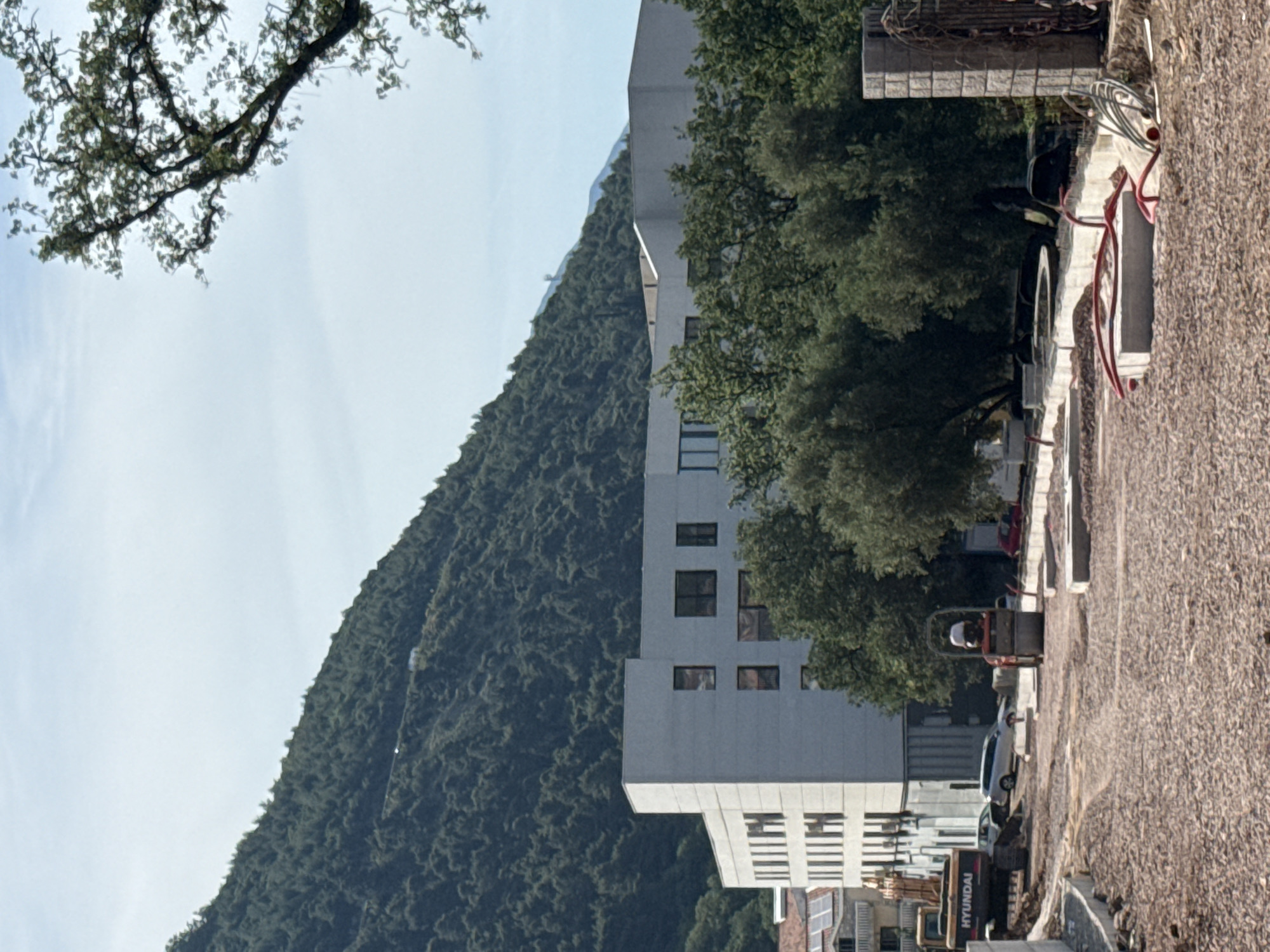 The image captures a scene near the city of Budva, Montenegro, featuring a stark contrast between natural and urban elements. Dominating the left side is a lush, green mountain covered in dense vegetation, while a modern, multi-story building with several windows is positioned on the right side. The building appears to be under construction or renovation, with construction vehicles and equipment visible in the foreground. A worker wearing a white hard hat is operating a piece of machinery. The foreground also features a section of a street or walkway covered in gravel, possibly due to ongoing construction. A branch from a tree extends into the frame from the top left corner. The sky is clear and light, indicating a sunny day.