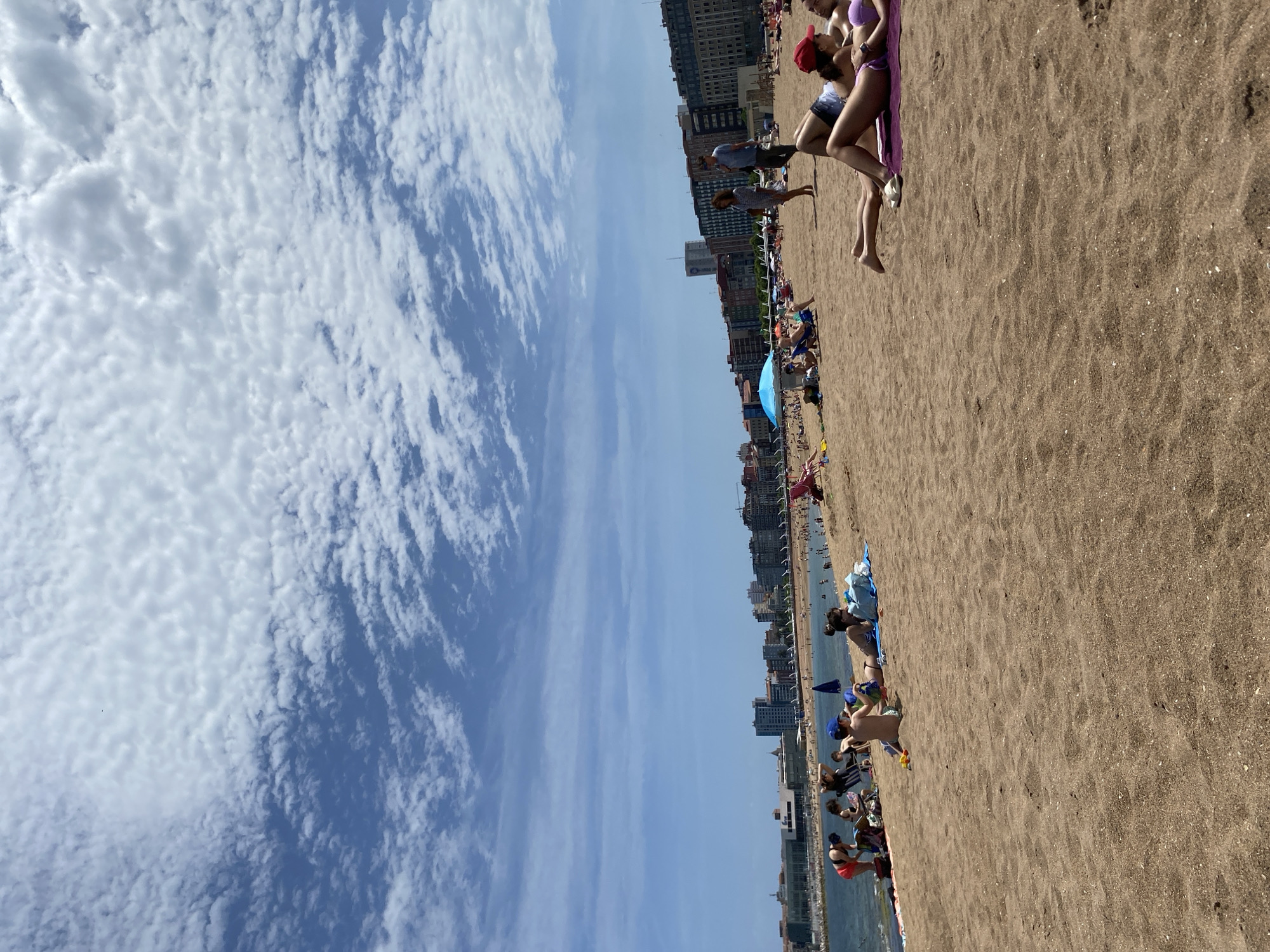 The image depicts a crowded beach in Gijón/Xixón, Spain, with people sunbathing and enjoying the day. The scene is dominated by a sandy beach with various individuals and groups scattered across it. Some people are lying on towels, others are seated, and some are standing or walking. There is a mix of clothing and beachwear visible, including swimwear, shorts, and shirts. Buildings line the background. The sky is partially covered with clouds.