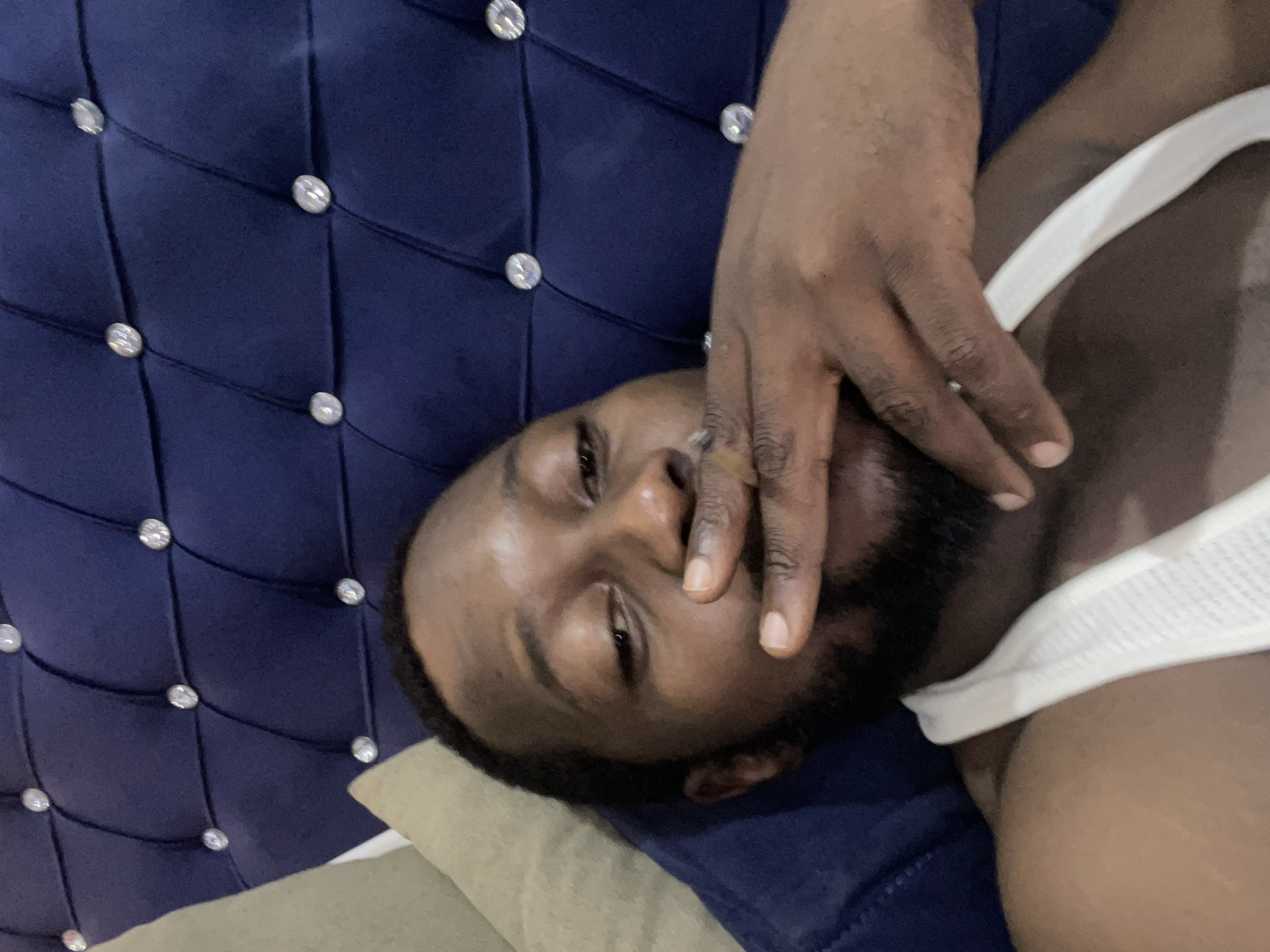 A dark-skinned man with a beard is lying on a bed. He is wearing a white tank top. His right hand is covering part of his face. The bed has a blue tufted headboard with diamond-shaped buttons. A pillow is beneath his head. The photo was taken in a place in the city of Abuja, Nigeria.