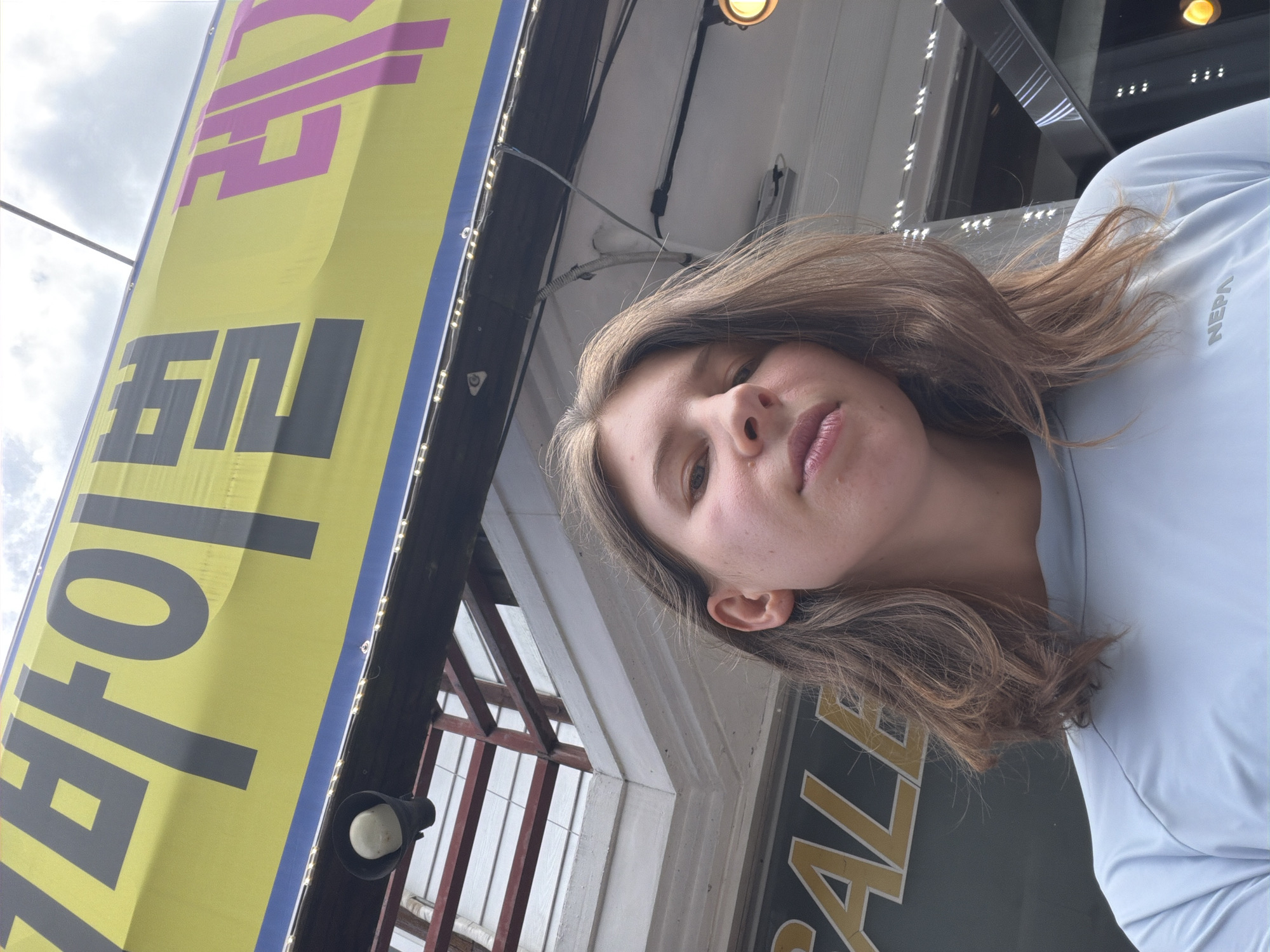 A young woman is pictured from a low angle, looking up and slightly to her left. She has long, light brown hair and is wearing a light blue t-shirt. Behind her, a large yellow banner with Korean text in black and pink is visible, as well as part of a building's exterior. The banner appears to be for a commercial establishment. A speaker is mounted on a dark beam above the woman. The overall scene suggests an outdoor setting, possibly a street or market area in Seo-myeon, South Korea.