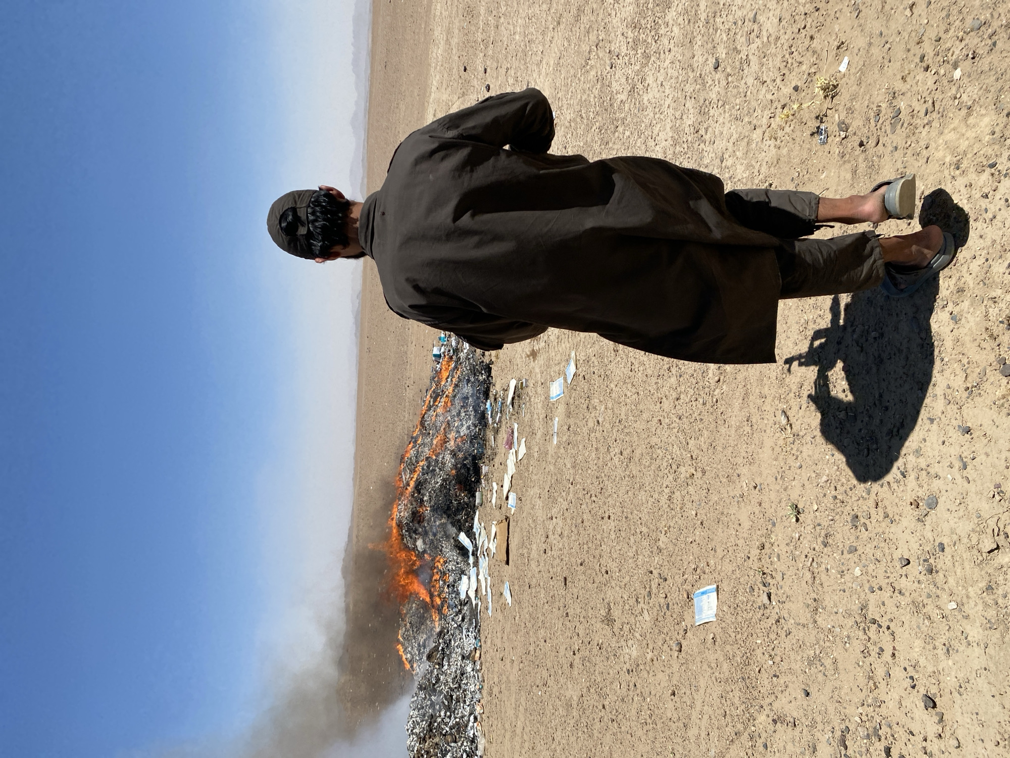 In the desolate landscape of Mandisar, Afghanistan, a solitary figure walks away from a large, smoldering pile of garbage. The fire, fueled by an unknown assortment of refuse, casts an orange glow against the dry, sandy terrain. Scattered across the ground are various discarded items, some appearing to be papers and what may be medical waste, such as face masks. The man, dressed in traditional dark clothing and a cap, is seen from behind, his shadow stretching long on the parched earth. The sky above is a clear, cloudless blue, emphasizing the vastness and emptiness of the surroundings. Distant mountains, hazy with dust, form a faint outline on the horizon, underscoring the remote and arid nature of the location.