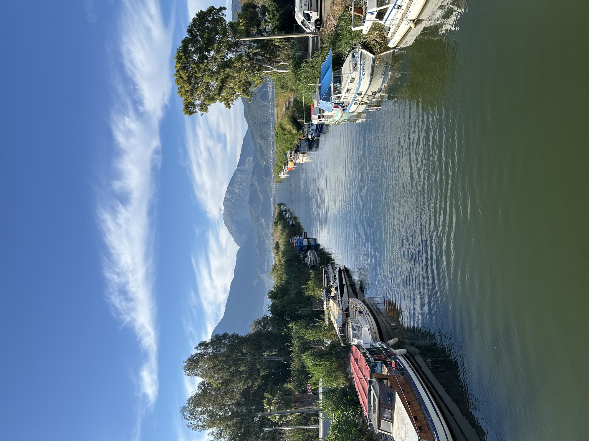 The scene is a canal or river bordered by moored boats on one side, and green vegetation and a mountain range on the other. The water is still, reflecting the sky and surrounding landscape. The sky is blue with wispy white clouds. The boats vary in size and condition. The mountain range is a prominent feature in the background, partially obscured by the wispy clouds. The location is likely the canals near Fethiye, Turkey, judging by the landscape and style of boats. No people are visible.