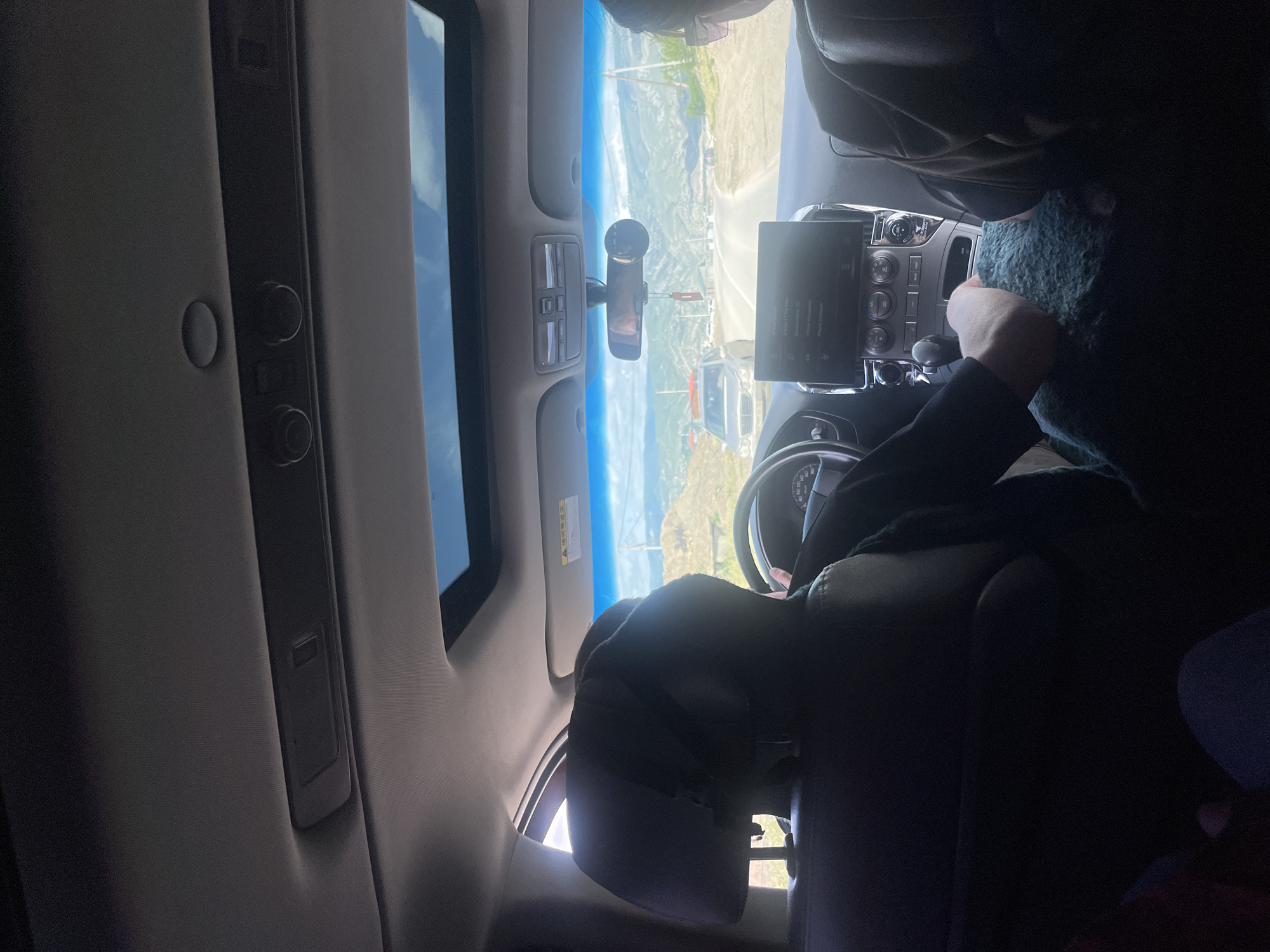 A person is in the driver's seat of a car, driving on a winding road. The location appears to be near the city of Chirkey, Russia, based on visible landscape features. A screen and dashboard are visible in the car's interior. The person is looking down and towards the right, possibly using a cloth or item held in their hand. The car has a sunroof.