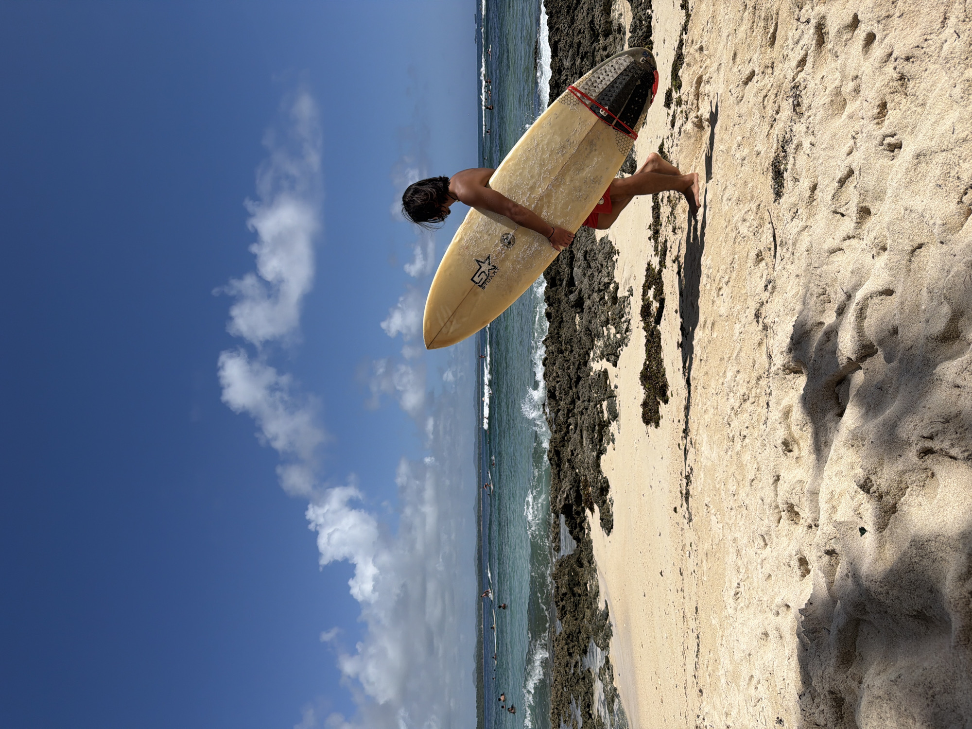 A person is holding a surfboard on a beach in General Luna, Philippines. The sky is blue with scattered clouds. The ocean has small waves, with people visible in the water. Rocky formations line the edge of the beach, and the sand is light in color. The person is fair-skinned, with dark hair. They are wearing red shorts and holding a yellow surfboard with a black and white patterned tail.