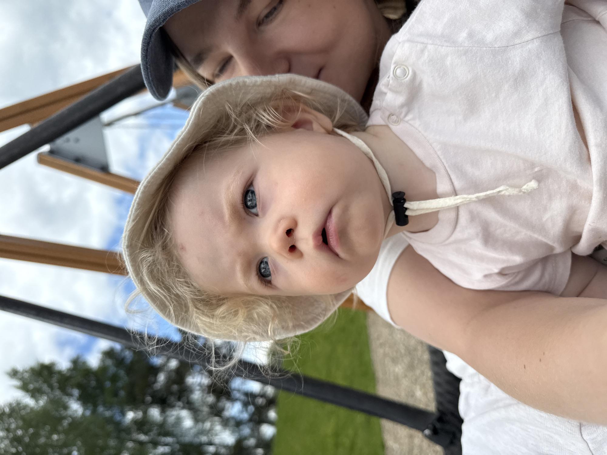 This close-up, rotated portrait captures an adult holding a young child outdoors in Dobrograd, Russia. The image is oriented sideways, giving the impression that the subjects are tilted.
The primary subject is a baby or toddler with fair skin, striking blue eyes, and blonde, curly hair. The child wears a cream-colored bucket hat with a chin strap secured by a black plastic cord stopper, and a light-colored, possibly pale pink, short-sleeved top featuring small buttons on the shoulder. The child looks directly at the camera with a curious expression and slightly open mouth.
Partially visible behind and above the child is an adult, likely female, wearing a dark blue or grey cap. Only the adult's forehead, eyes, and part of their cheek are discernible, suggesting a gentle, smiling expression as they hold the child close.
The background is softly blurred but reveals elements of an outdoor environment under a bright, cloudy sky, indicating daytime. Visible cues include brown wooden beams and black metal structures, along with hints of green grass and trees, suggesting a park or playground setting. There is no visible text in the image. The scene conveys a tender and affectionate moment between the adult and child during an outing.