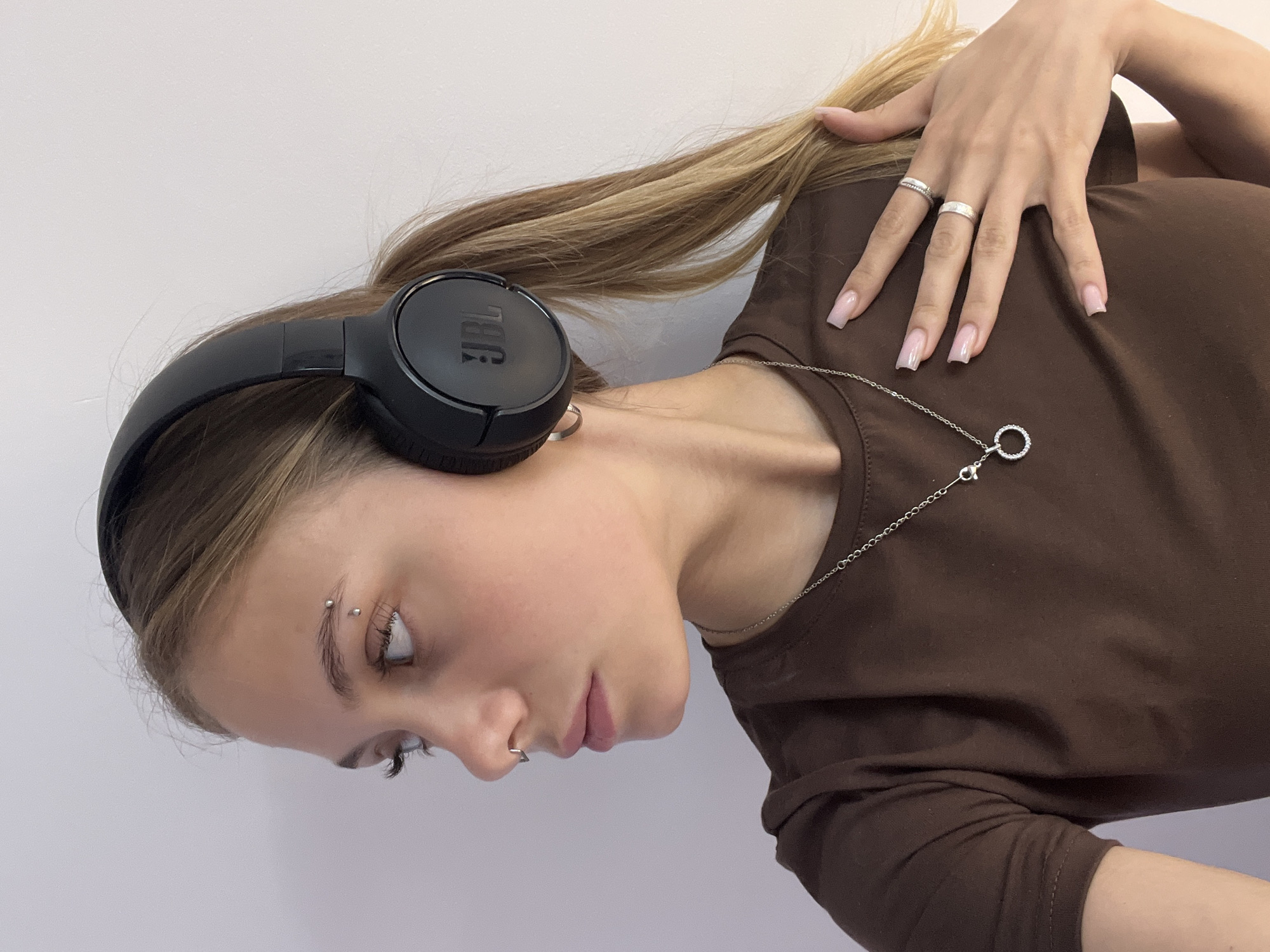 A young woman with light brown hair and light skin is depicted from a high angle. She is wearing black JBL headphones and a brown long-sleeved shirt. She has multiple piercings, including one in her eyebrow and one in her nostril. She is wearing a silver necklace and two silver rings on her hand, which is resting near her neck. The background is a plain, light gray wall. The location in the city of Фоки, Russia cannot be determined from the visual information.