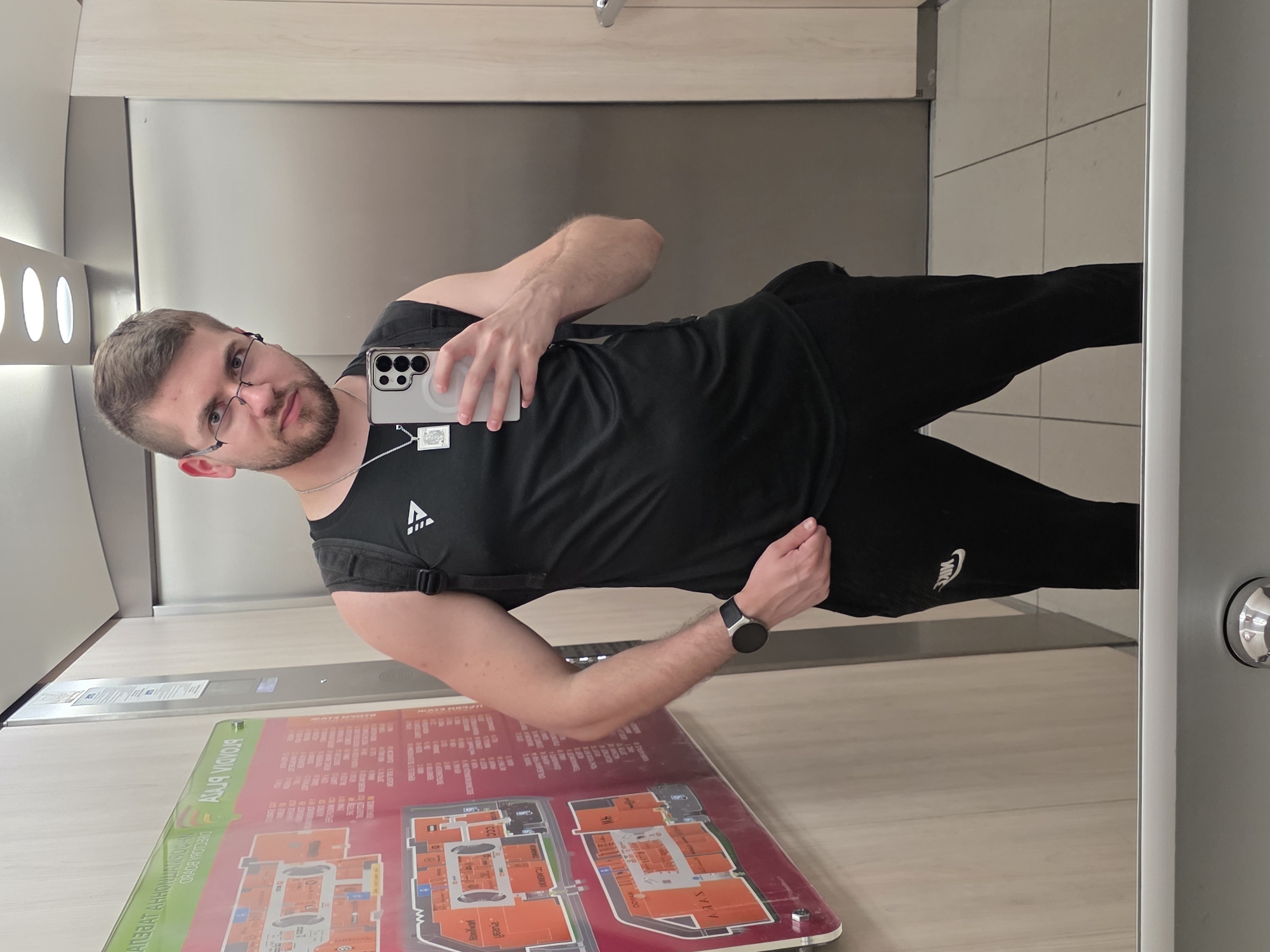 A man is taking a selfie in what appears to be an elevator. He has short brown hair, a beard, and glasses. He is wearing a black tank top with a logo on the chest and black Nike sweatpants. He's holding a phone with a gray case. In the elevator, there's a directory map, suggesting the location is in a shopping center. The map's text includes what looks like Bulgarian language, and based on the Cyrillic script, the word "Пловдив" can be clearly identified. This indicates the shopping center is located in Plovdiv, Bulgaria. The elevator has a mirrored wall reflecting the man and the map. The floor tiles are light beige. The elevator has built-in, long, and narrow light fixtures in the ceiling.