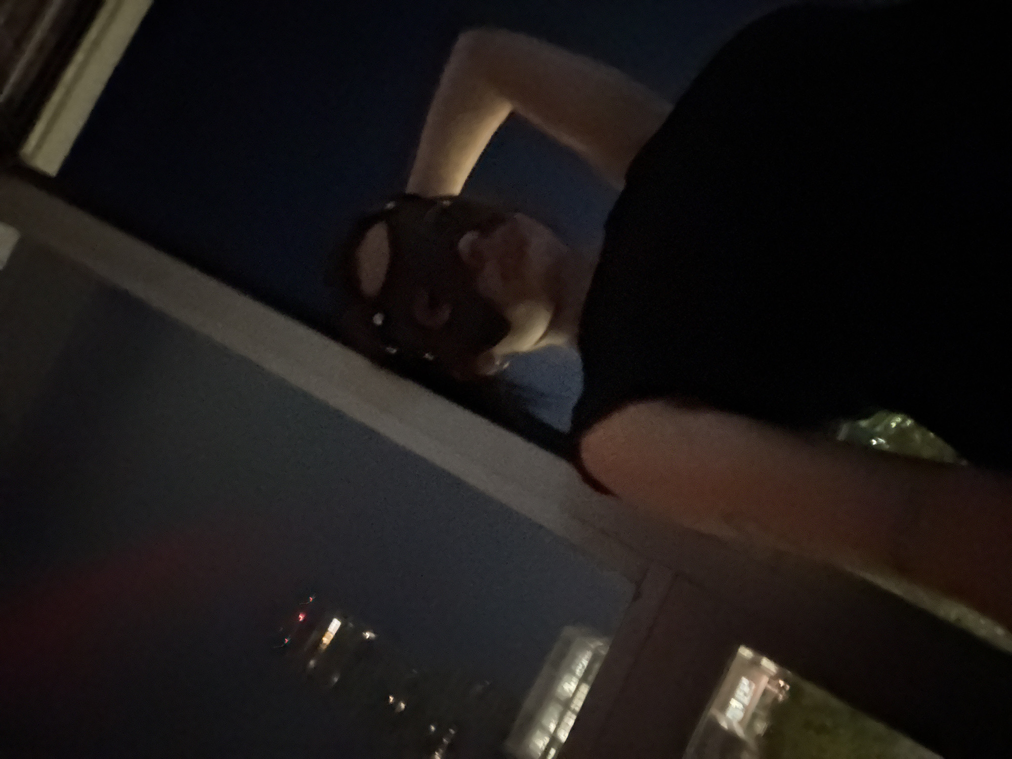 The image captures a person leaning out of a window at night, looking down or out at a city scene, likely in Buenos Aires, Argentina. The photograph is taken from a low angle, looking upwards, with a prominent window frame and sill in the foreground and midground.
The main subject is a person, visible from the chest upwards. They are wearing a dark-colored t-shirt. Their left arm is bent, with the elbow seemingly resting on the windowsill. Their head is tilted downwards and to the side, and a dark, possibly reddish-brown accessory or hat is on their head, obscuring some facial details. Their skin is light-toned.
The scene outside the window is a dark night sky, indicating the time of day is evening or night. The background features blurred, distant city lights, suggesting a dense urban environment. These lights are visible as small, bright specks and streaks, primarily in yellow, white, and red hues, against the dark expanse. Some illuminated building facades are vaguely discernable in the distance.
The activity depicted is the person observing the nocturnal cityscape from an elevated vantage point. The image conveys a sense of quiet observation or contemplation. No specific weather conditions are discernible, and no visible text is present.