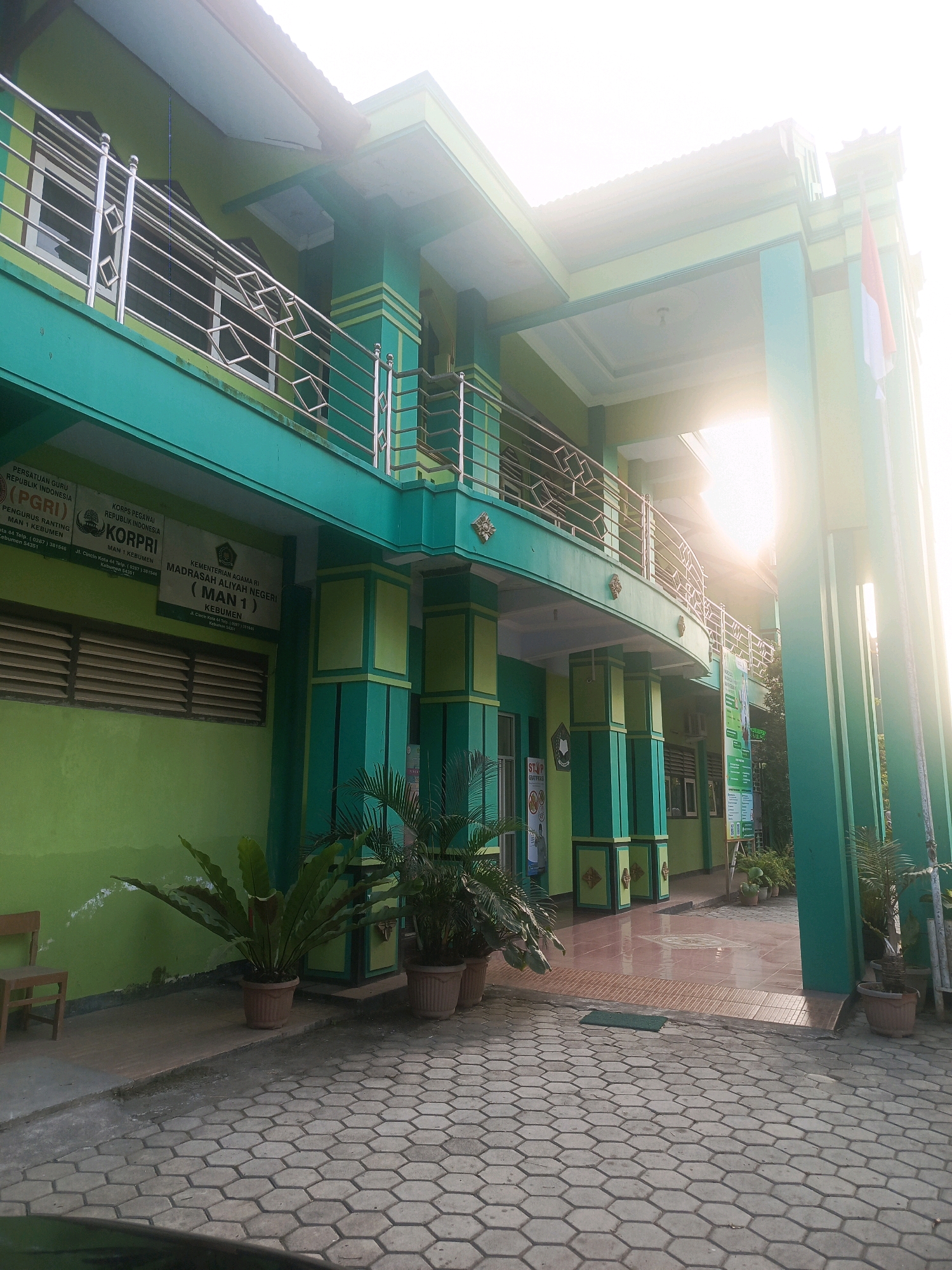 The image depicts the exterior of a two-story building, painted in vibrant green and yellow hues, with metal railings on the balconies. The building appears to be a school or administrative facility. Several potted plants, including ferns and palm-like varieties, are placed on a paved walkway in front of the building. A small wooden chair is visible to the left, near a green wall. The sky is bright and somewhat overcast, with sunlight filtering through, casting a bright glow on the upper parts of the building and creating lens flare effects. The ground is paved with hexagonal tiles. There are signs on the building's facade, one with an Indonesian flag and others with text that is partially legible, suggesting it is a public institution in Indonesia. Based on the text on the signs, which includes "MAN 1 Kebumen" and "KORPRI," the location is likely Madrasah Aliyah Negeri 1 Kebumen, a public religious high school in Kebumen, Central Java, Indonesia. There are no people visible in the image.