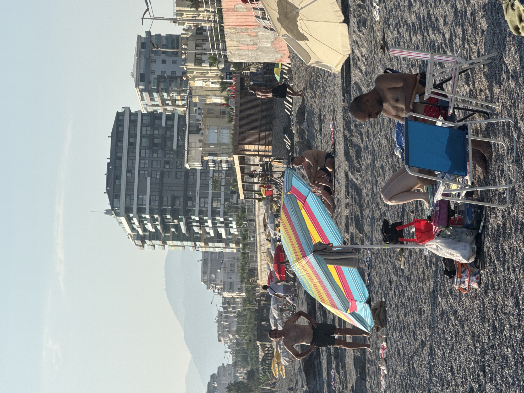 A beach scene in Alanya, Turkey, on a bright, clear day. In the foreground, a man sits on a folding chair, looking down and appearing to be engaged with something in his hands. Next to him, another empty blue folding chair is angled towards him. Behind this pair, a colorful beach umbrella is set up. Further back on the beach, several people are seated or standing, with more umbrellas and beach chairs scattered across the pebbly sand. In the middle ground, a man in dark shorts stands with his hands on his hips, facing the camera. Beyond the beach, a row of multi-story buildings, some with balconies, forms a cityscape. In the distance, hazy mountains rise against a pale blue sky. The scene depicts a typical day at a tourist beach, with people enjoying leisure time under the sun.