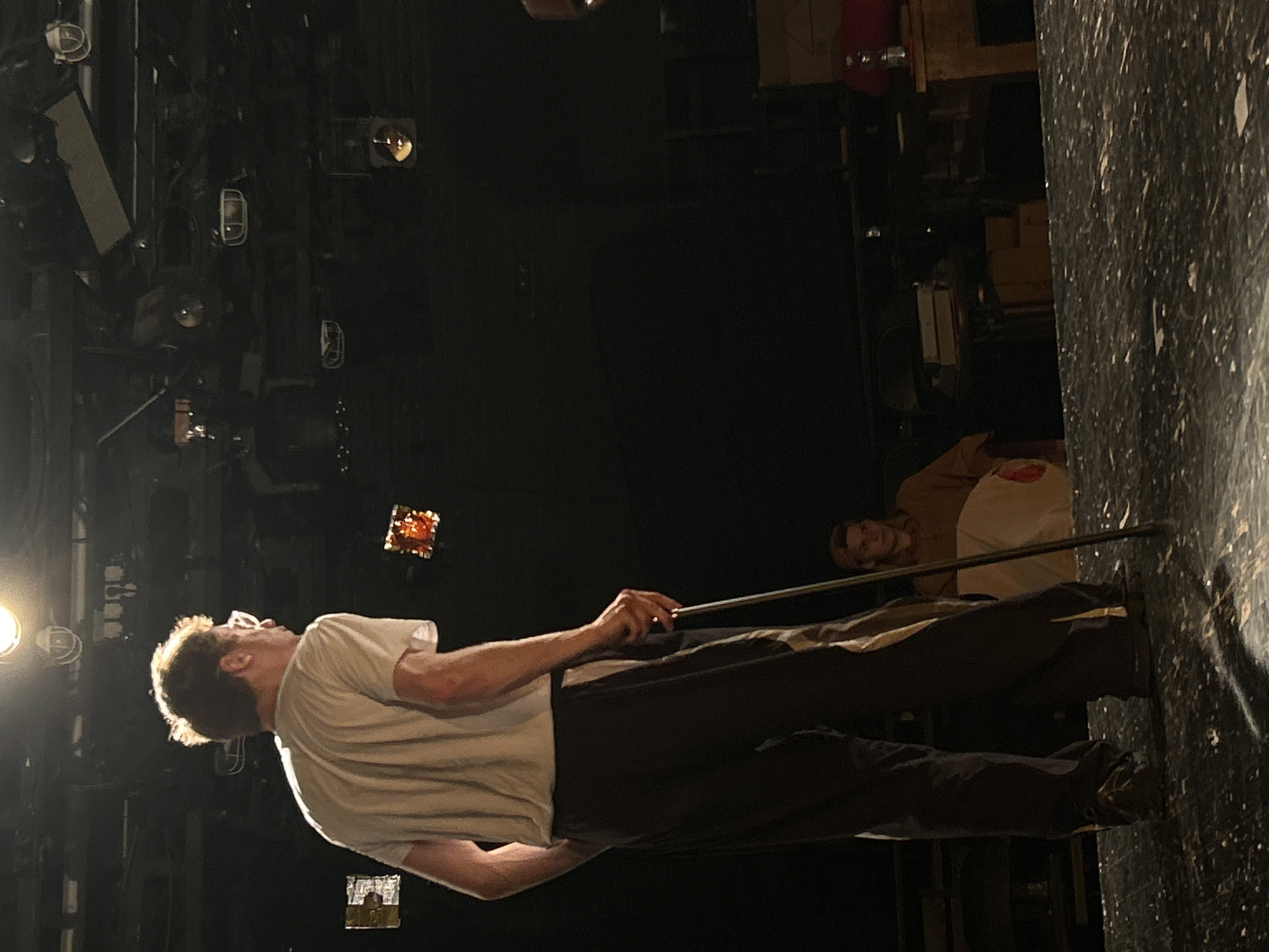 A young man, wearing glasses and a white t-shirt, stands on a stage holding a cane, with his back to the camera. He is wearing dark pants with a stripe down the side. In the background, another person wearing a brown hoodie and a baseball cap is seated. The stage appears to be dimly lit with spotlights from above. The floor of the stage is textured and dark, with some debris. The overall setting suggests a theater or performance space. The location is Unknown, Unknown.