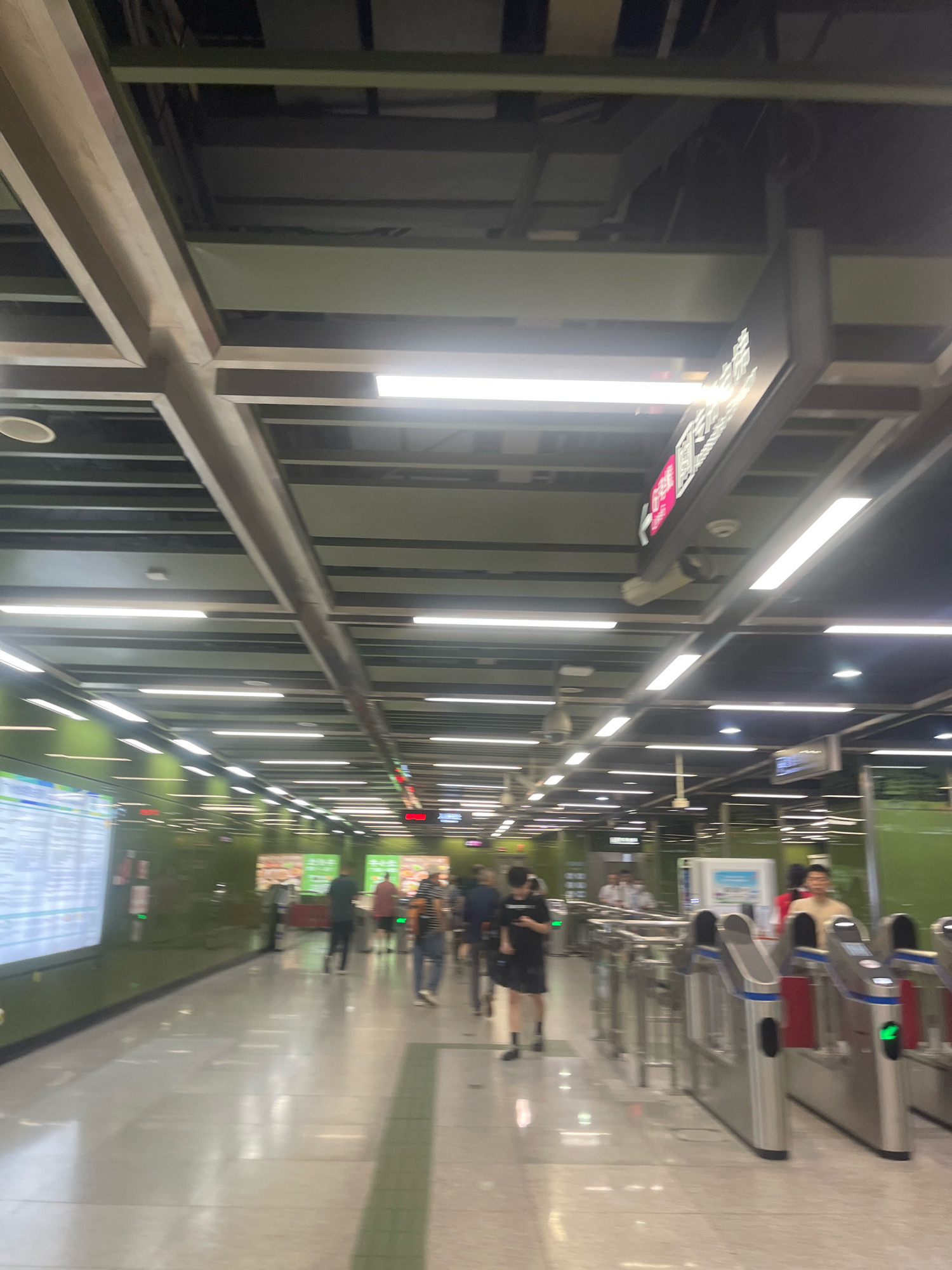 This is a photograph of a subway station concourse in Yuexiu District, China. The ceiling is high, with exposed ductwork and rows of fluorescent lights. The walls on the left are lined with informational displays and green panels, while the right side features ticket gates and more promotional signage. Several people are visible walking through the station, some appearing to be passing through the ticket gates. The overall impression is of a modern, well-lit, and functional public transportation hub.