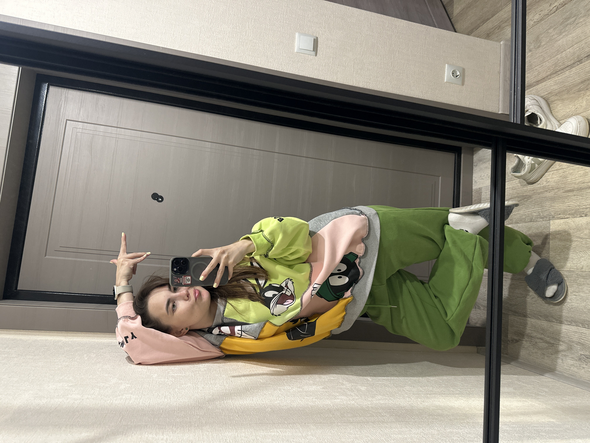 A young woman is taking a selfie in a hallway. She is wearing a colorful hoodie with Looney Tunes characters and green sweatpants. She is leaning against a wall with her arm raised, holding her phone to take the photo. Her reflection is visible in a large mirror that takes up the right side of the frame. In the background, a door and a light switch are visible. The floor is made of light-colored wood. The scene appears to be an indoor residential setting.