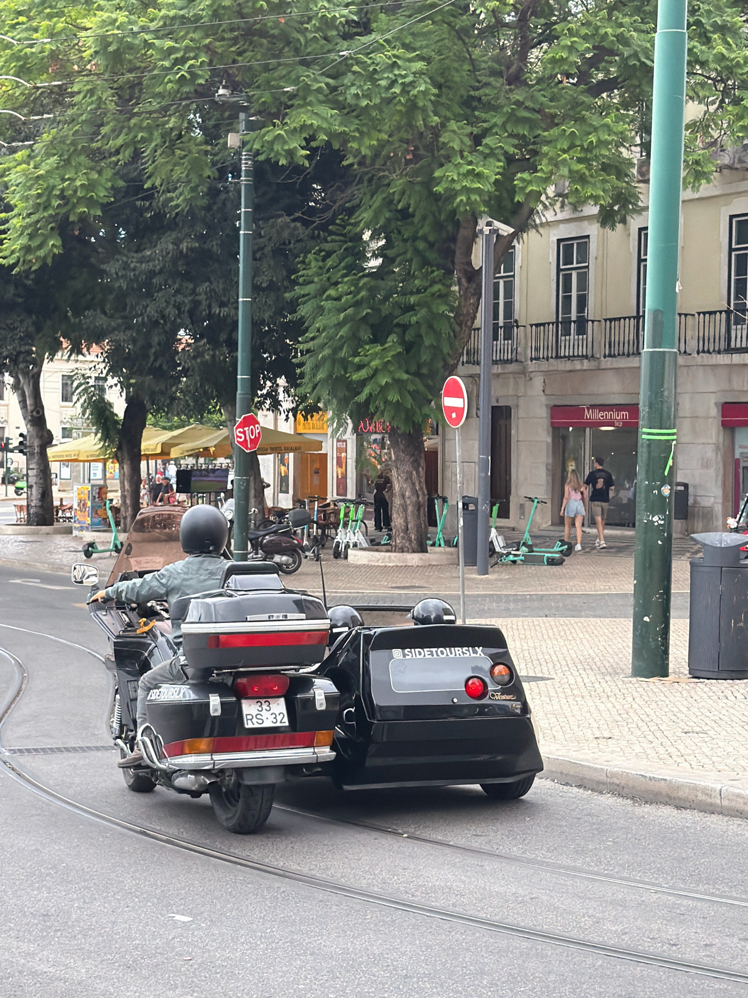 In Lisbon, Portugal, a motorcycle with a sidecar is traveling on a tram track on an overcast day. The motorcycle is black with a red taillight and a license plate that reads "33 RS 32". The sidecar also has a logo that says "SIDETOURSLX". The motorcycle is being ridden by a person wearing a gray jacket and a black helmet. In the background, there are buildings, trees, and a stop sign. There are also several electric scooters parked on the sidewalk. The scene appears to be in a tourist area, with shops and cafes visible.
