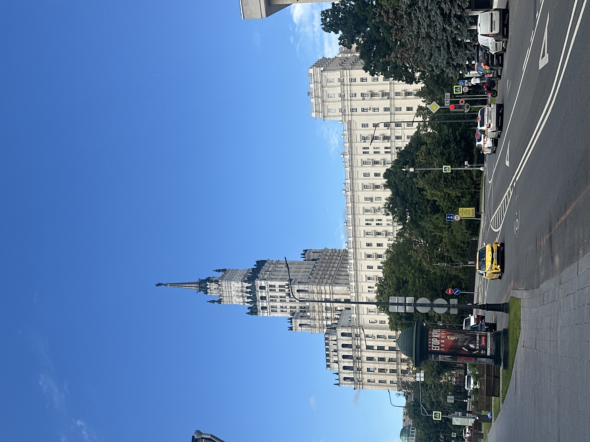 This is a vertical, medium-distance shot of the Kotelnicheskaya Embankment Building, a prominent Stalinist skyscraper in Moscow, Russia. The building is made of light-colored stone and features a central, ornate spire topped with a star. The skyscraper is flanked by two lower wings that extend outward. Trees and a street are visible in the foreground, with cars and pedestrians on the road. A yellow taxi is seen driving on the right side of the street. The sky is clear blue with a few wispy clouds. A large poster is displayed on a cylindrical stand on the left side of the sidewalk, advertising a performance. Streetlights, traffic signs, and crosswalks are also visible. The overall scene depicts a sunny day in a busy urban environment.