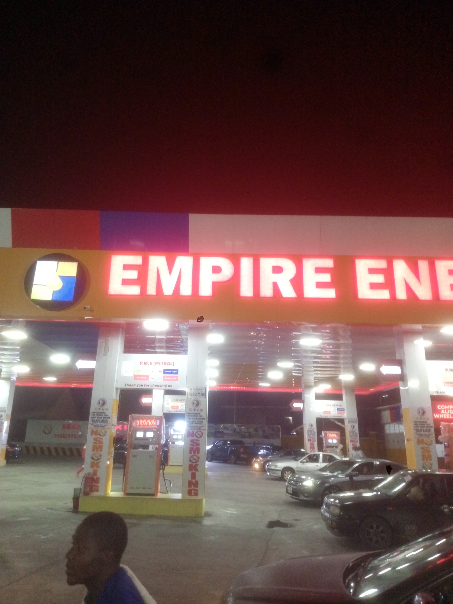 The image shows an "Empire Energy" gas station at night in Gwarinpa, Nigeria. The gas station's sign is illuminated, and several cars are parked or driving at the pumps. A man is visible in the lower left corner. "No Smoking" signs are posted near the pumps. P.M.S (Petrol) service is visible, and a sign offers a phone number for complaints. The ground is paved and shows signs of wear.