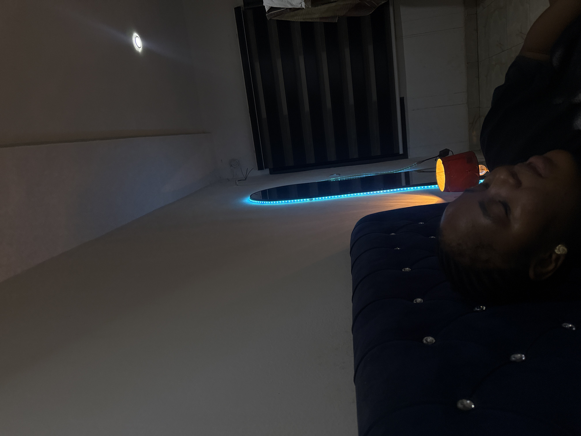 The image depicts a person reclining on a tufted, dark-blue upholstered surface. They are wearing dark clothing. The scene is indoors, with a partial view of a window covered by dark vertical blinds. A round table with blue LED lighting around its perimeter sits near the window. An orange cylindrical lamp is also present near the table. Based on the interior design elements, including the tufted upholstery, the LED lighting, and the design of the window blinds, it is likely that the location is in Abuja, Nigeria, a city known for its modern architecture and interior design trends.