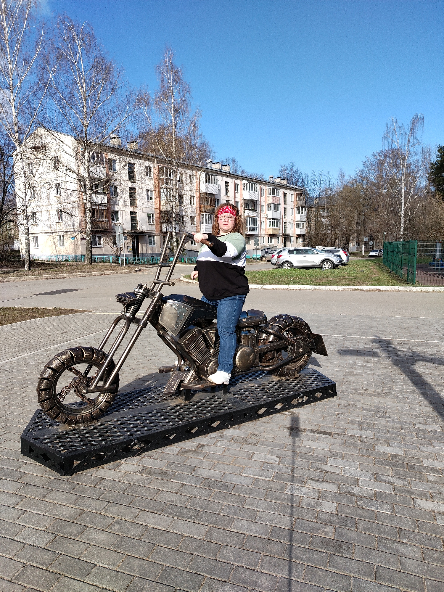 The image shows a person sitting on a metal sculpture of a motorcycle. The person is wearing a red bandana, glasses, a long-sleeved striped shirt, blue jeans, and white shoes. The sculpture is positioned on a black metal platform, which sits on a paved area of gray bricks.
In the background, there is a multi-story building with beige walls and multiple windows, typical of Soviet-era architecture. Several cars are parked in a grassy area behind the sculpture. Bare trees and a blue sky with light clouds are visible in the background.
The location is Chaikovsky, Russia. The image captures a scene of a person interacting with a piece of public art in a city environment.