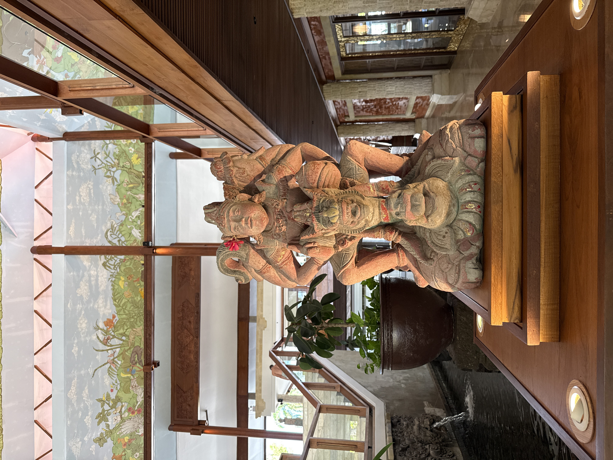This image displays an ornate indoor architectural space, likely in Kuta Selatan, Indonesia, featuring traditional design elements and a prominent statue. The central subject is a large, intricately carved statue, possibly made of stone or wood, depicting figures that appear to be from Hindu mythology. The statue shows signs of weathering and hints of red, gold, and grey paint, portraying a multi-armed deity figure atop what looks like a mythical creature.
The statue is positioned on a polished wooden console, which is illuminated by circular recessed lights and overlooks a dark water feature, possibly an indoor pond or pool. Behind the statue, a large potted plant adds a touch of greenery.
The surrounding environment is characterized by rich wooden architecture. Above, a ceiling with dark wooden beams and slatted panels is visible. To the left, decorative panels, resembling murals or intricate screens behind glass, depict lush natural scenes. These panels feature vibrant foliage, flowers, and various animals, including birds, in a palette of green, light blue, and white. Further into the background, additional architectural elements such as textured stone walls and windows suggest a spacious, multi-level building. The scene evokes a sense of cultural richness and tranquility, possibly a hotel lobby or a large traditional building, captured during daytime. No text is visible in the image.