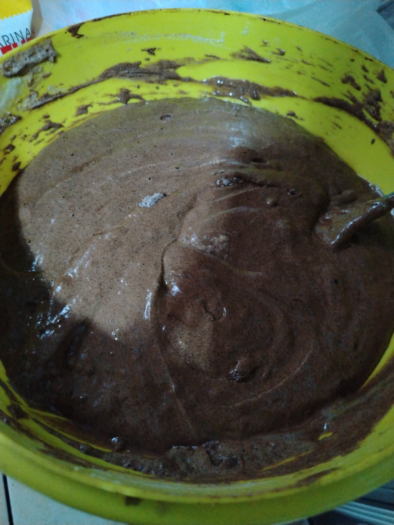 A close-up, eye-level shot of a yellow mixing bowl filled with dark brown batter, likely cake or brownie batter. The batter has a slightly shiny appearance and is somewhat uneven in texture, indicating it has recently been mixed. The bowl itself has batter residue smeared on its sides. The top left corner of the image shows a partial view of a package, possibly flour, with the word "RINA" visible.
There is no information in the image to verify its location in Tinaquillo, Venezuela, or provide specific details about people, actions, or interactions beyond the batter and its container.