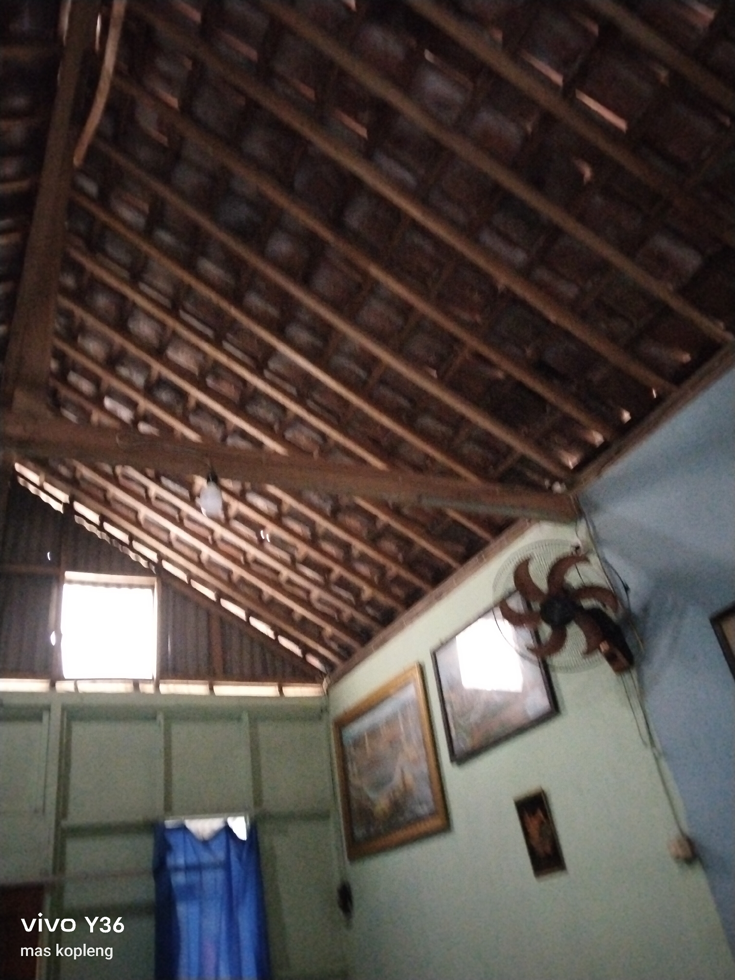 The image shows the interior of a rustic house, likely in Baturetno, Indonesia, with an exposed wooden roof structure and traditional tile roofing visible from below. The walls are a pale green, and a blue curtain hangs behind a wooden partition. On the wall, there are several framed pictures, including one larger landscape painting and a smaller, darker framed image. A vintage-style fan is mounted on the wall to the right of the paintings. A light bulb hangs from the ceiling. The lighting suggests it is daytime, with light entering from a window visible on the left. The overall impression is of a simple, lived-in space.