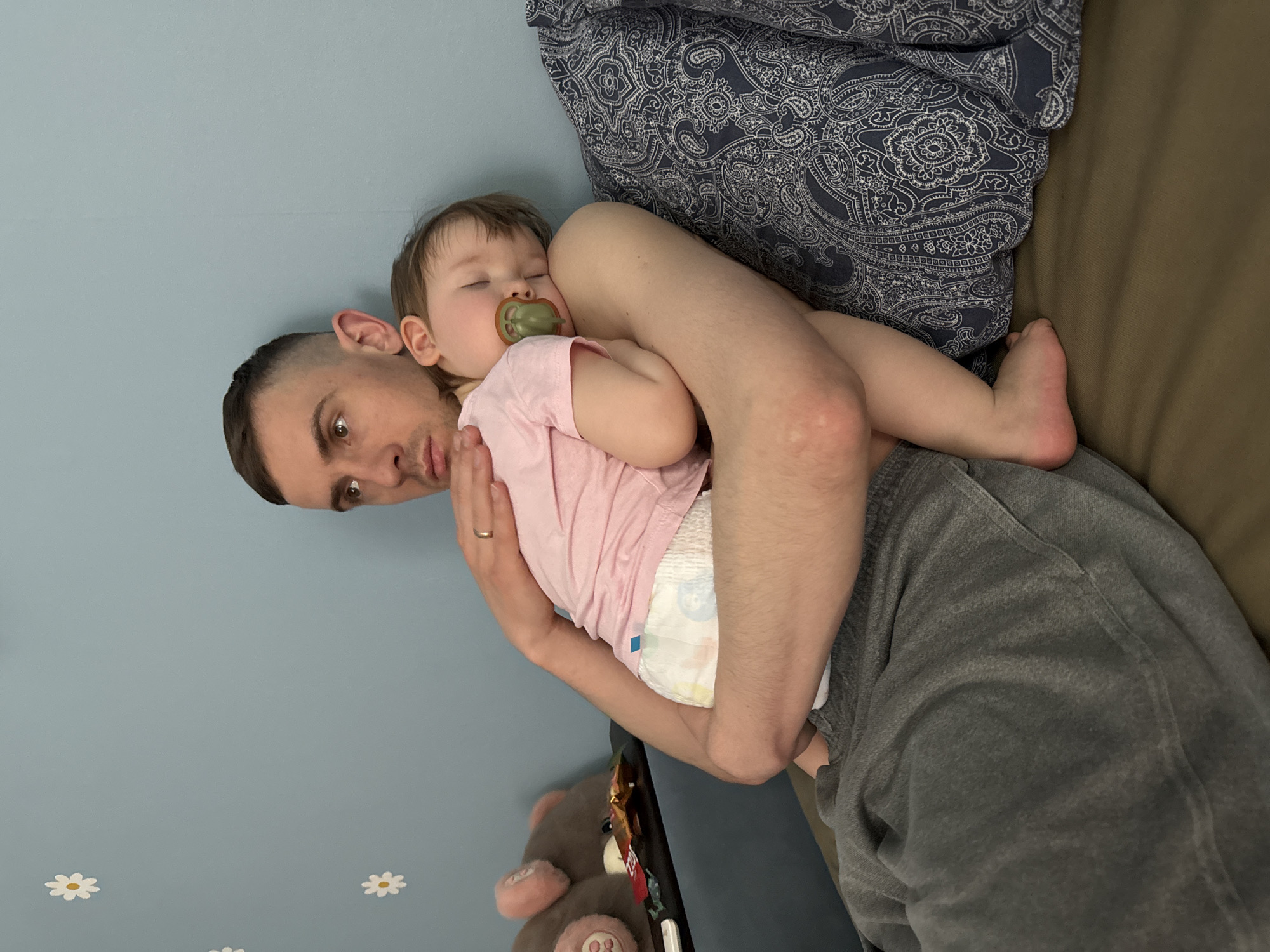 A man is holding a sleeping baby in his arms. The baby has a pacifier in its mouth and is wearing a diaper and a pink shirt. The man is wearing gray sweatpants and a wedding ring on his left hand. The background shows a blue wall with white daisy stickers, a patterned blanket, and a brown couch. The location is not discernible and cannot be determined to be Moscow, Russia.
