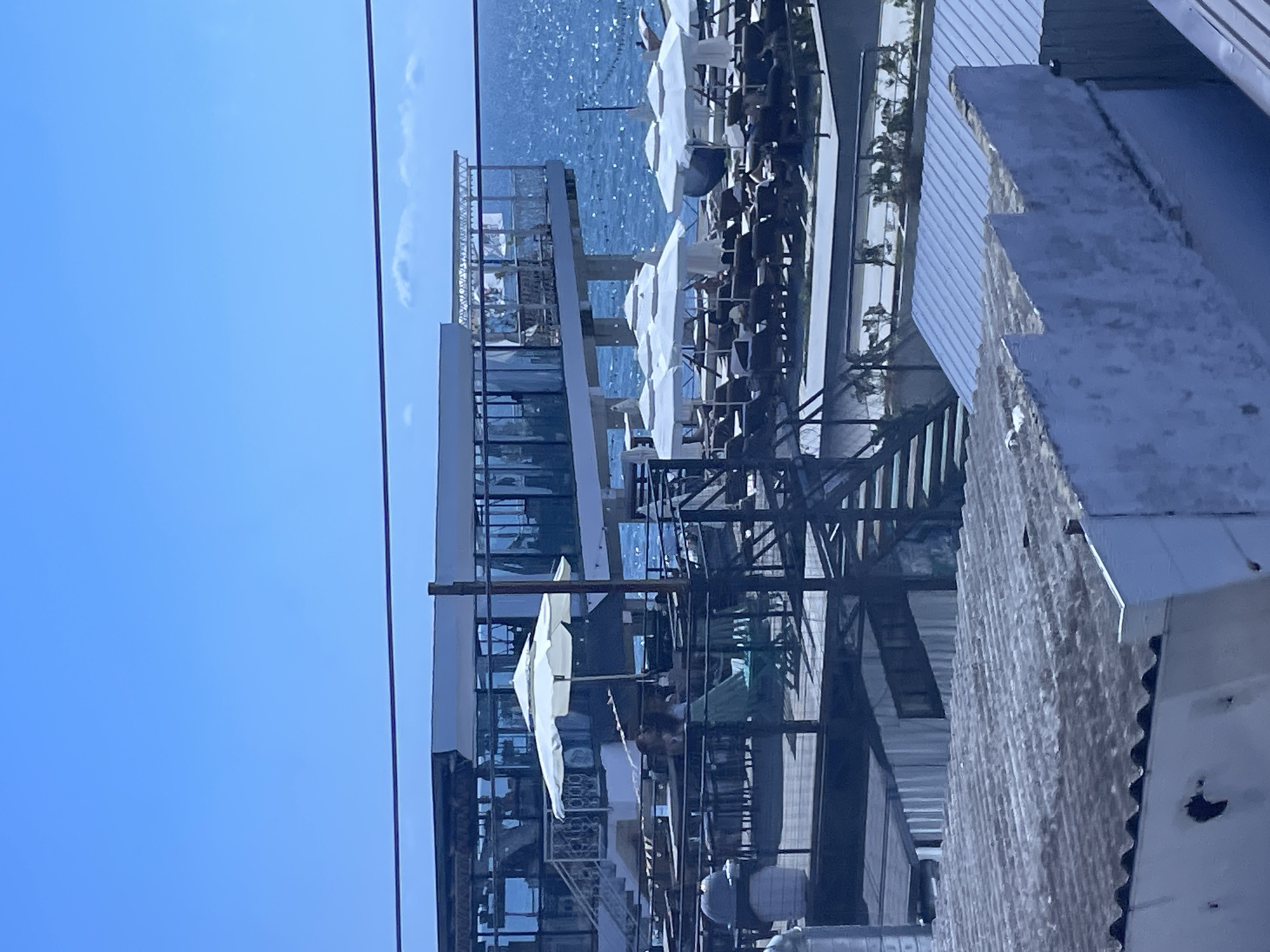 The image depicts a resort setting on a sunny day, likely in Sochi, Russia, given the presence of a modern building structure and the typical ambiance of a seaside resort. In the foreground, the corrugated metal roofs of two low-rise structures dominate the lower portion of the frame. The structure on the left appears to be weathered with some discoloration and a hole visible. The structure on the right has a clean, light blue corrugated metal roof.
Behind these roofs, a multi-level outdoor seating area is visible. Tables and chairs are arranged on a wooden deck, with a white umbrella providing shade for two individuals seated at a table. One person is wearing a light green dress. Further back, a wider area is set up with rows of lounge chairs, many of which are covered by white umbrellas, suggesting a beach or pool area.
The background is dominated by a modern, multi-story building with extensive glass windows, suggesting a hotel, restaurant, or other establishment. The building is elevated on pillars and overlooks the sea. The sea itself is a vibrant blue, sparkling under the sunlight, with some gentle waves. A thin, dark cable runs horizontally across the upper part of the image, cutting across the sky. The sky is clear and bright blue, with a few faint clouds visible on the horizon. The overall impression is one of a leisure destination, with a clear division between the functional, industrial-looking foreground structures and the more elegant resort facilities in the background.