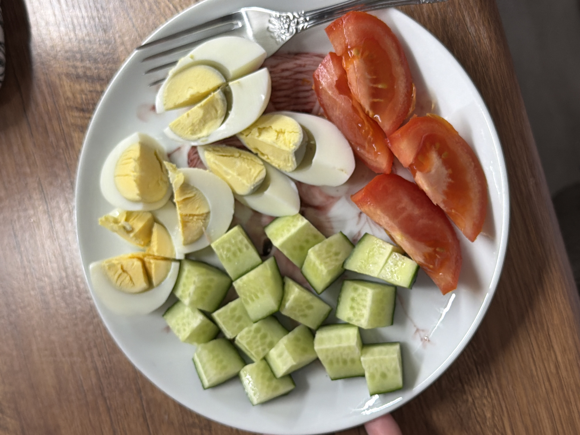 An overhead close-up shot captures a white plate filled with a fresh and healthy meal, resting on a brown wooden surface, likely a table.
The plate features an arrangement of simple, wholesome ingredients. On the left side, several hard-boiled eggs are neatly cut, with some halved and others quartered, clearly displaying their firm, bright yellow yolks and white albumen. Adjacent to the eggs, towards the bottom-center of the plate, is a generous portion of diced green cucumbers, cut into small, uniform cubes. On the right side of the plate, several vibrant red tomato slices, some appearing slightly wedge-shaped, are arranged.
A silver fork with an intricately designed handle is partially visible at the top edge of the plate, indicating the meal is ready to be consumed. The plate itself is white with a very subtle, almost translucent, pattern. The wooden table surface below has a distinct grain pattern, suggesting an indoor setting.
The image depicts a prepared meal, possibly a light breakfast, lunch, or snack, characterized by its fresh, unadorned ingredients. There is no visible text in the image.